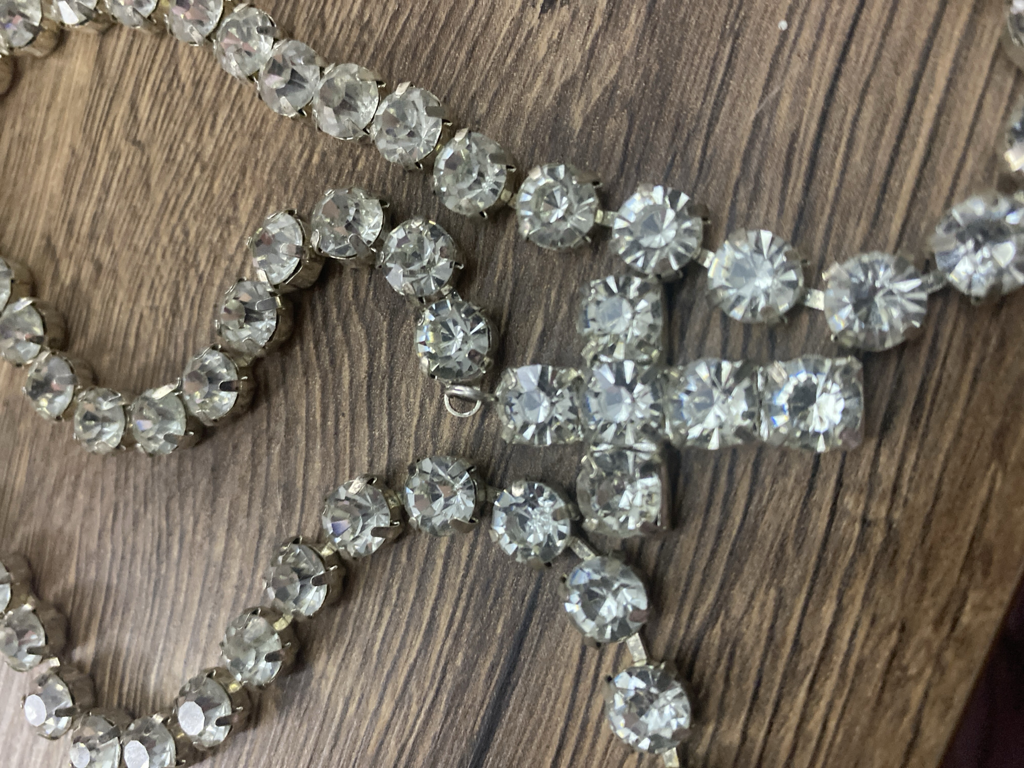 A close-up, overhead shot shows a sparkling, silver-toned necklace with clear, faceted stones lying on a dark wood grain surface. The necklace is made up of numerous round and square-shaped stones, each set in a metallic prong setting. The stones catch the light, creating a dazzling effect. The necklace appears to be intertwined and layered in some areas, suggesting it might be a bracelet or a long pendant necklace. The wood surface has visible grain patterns and appears to be a table or countertop. The overall impression is one of glitz and glamour, with the necklace as the central focus.