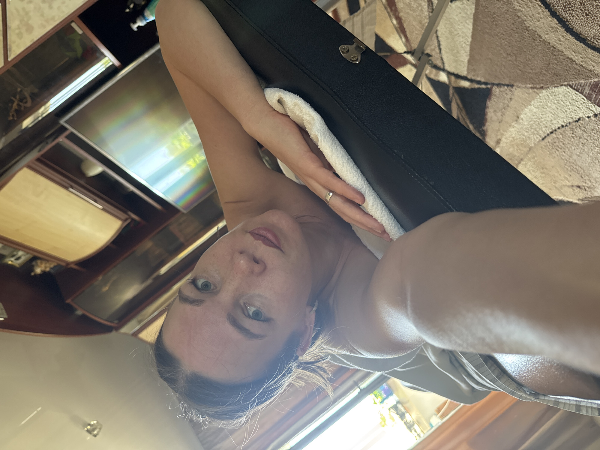 A woman lies on her back on a black, padded surface, possibly a massage table or similar piece of furniture. She has light skin, light eyes, and her hair is pulled back. Her arms are crossed above her head, and she is wearing a ring on her left hand. A white towel is positioned between her head and the black surface. The background includes a display cabinet with glass doors and shelves, suggesting an interior setting. A patterned rug is visible. Based on available information, the location is Chaikovsky, Russia. The image is a self-portrait or taken from a low angle.