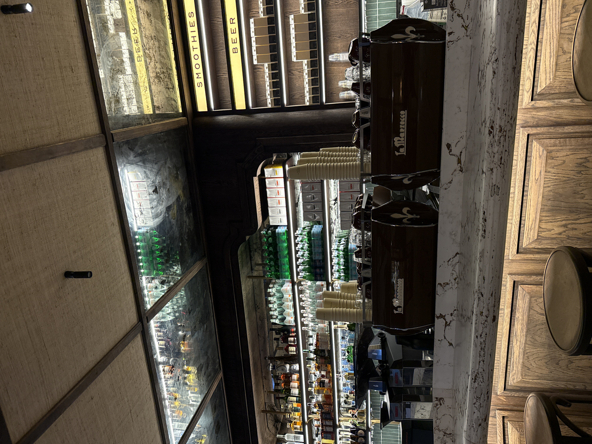 The image shows the interior of a bar. A counter with a marble top runs from left to right, with stools next to it. Behind the counter, there are shelves stocked with bottles, likely alcoholic beverages. There is an espresso machine, cups, and a sign that says "Smoothies" and "Beer." The scene appears to be indoors, in a dimly lit setting. The location could be in Cape Town, South Africa, based on the type of establishment and possibly the available beverages. There are no people visible in the image.