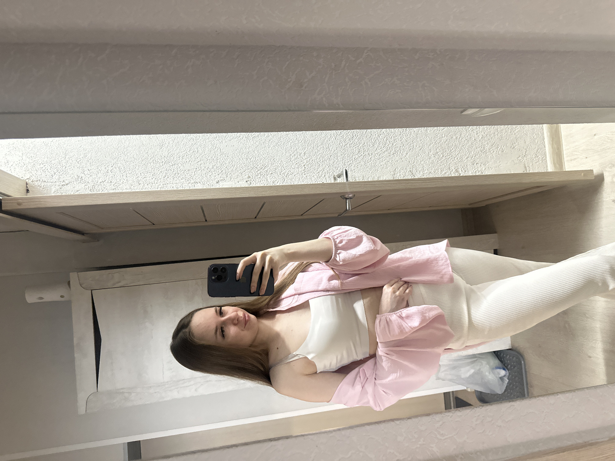 A mirror selfie captures a young woman standing indoors. She has long, straight brown hair, which falls past her shoulders, and is looking towards the camera with a gentle smile. She is holding a dark-colored smartphone, likely an iPhone due to the visible camera module, in her left hand to take the picture. She is dressed in a white sleeveless crop top, layered with a light pink, long-sleeved overshirt or jacket made of a crinkled fabric. Her lower body is visible, wearing light-colored, possibly off-white, ribbed pants.
The background shows a brightly lit room with textured, light-colored walls. Above the woman, there's a light-colored wooden or laminate shelf or cabinet. On the underside of this shelf, small metal hooks are visible. To the left, on another shelf or surface, a white cylindrical dispenser can be seen. Below the woman, a light-colored floor is visible, with a small gray mat and a crumpled plastic bag in the bottom right portion of the frame. The overall setting suggests a casual home environment, possibly a utility area or dressing room. There is no visible text in the image.