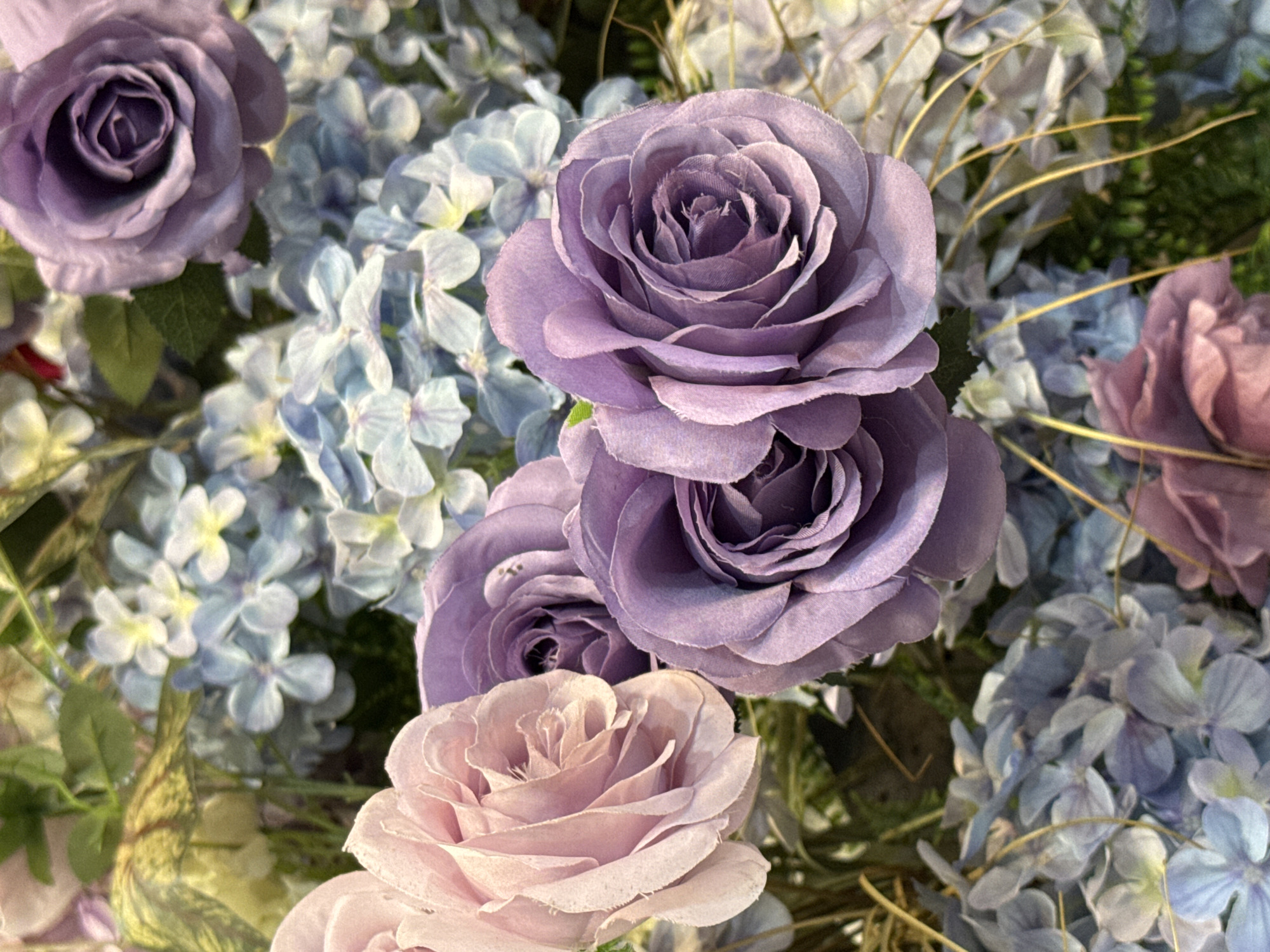 The image displays an arrangement of various flowers, predominantly roses and hydrangeas, in shades of purple, lavender, and light pink. The roses exhibit a soft, velvety texture with tightly spiraled petals forming the central part of the blooms. The hydrangeas are grouped in clusters, with small, individual florets creating a fuller, textured appearance. Thin, straw-like elements are interspersed among the flowers. The image does not contain information to determine its specific location as Qianjiang, China, or any information regarding people, actions, or interactions.