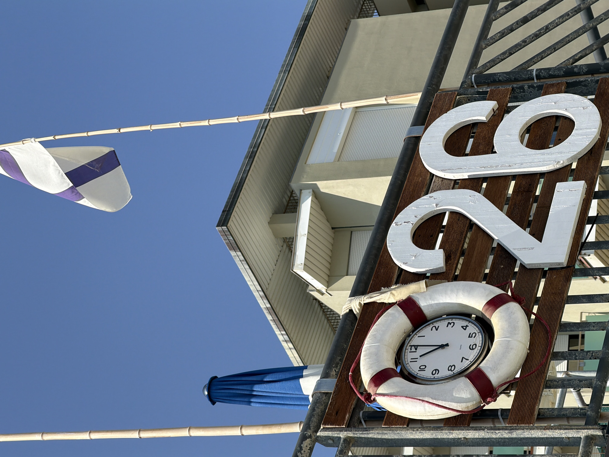 The image shows a close-up of a sign with the number "26" in large white numerals against a wooden plank background. To the left of the numerals, a white life preserver with red stripes is hung, and inside it, a circular clock face is visible. The clock hands indicate it is approximately 10:00. Behind the sign, a building with a light-colored facade and a sloped roof is visible. A blue and white umbrella is folded and leaning against a pole to the left of the building. A flag with purple and white stripes is flying from a bamboo pole above the building. The sky is clear and blue. This appears to be a beachside location in Gatteo, Italy, likely at a beach establishment or bathing establishment numbered 26. There are no people visible in the image.