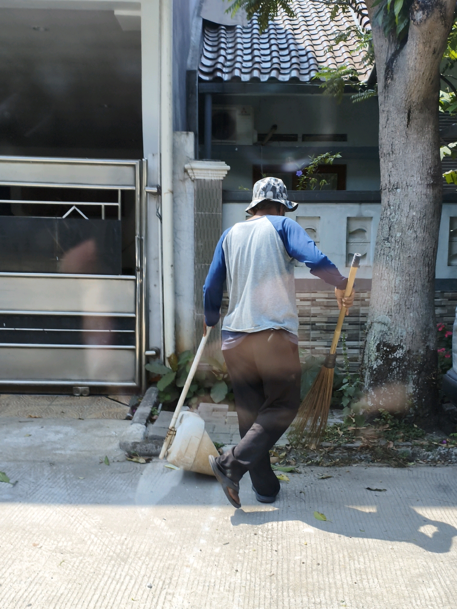 A person wearing a blue and white long-sleeved shirt and a camouflage bucket hat is sweeping the ground with a broom and a dustpan. The person is walking away from the camera, with their back mostly visible. They are wearing dark pants and sandals. The broom has a bamboo handle and a straw brush. The dustpan is white and cone-shaped. The ground is paved with concrete and has a patterned texture. In the background, there is a house with a tiled roof and a large tree. The scene appears to be on a street or sidewalk in Sukaraja, Indonesia. There is a metal gate to the left of the frame, which appears to be part of the house's property. Sunlight is casting shadows on the ground, suggesting it is a bright, sunny day. Green foliage is visible around the base of the tree and near the house.