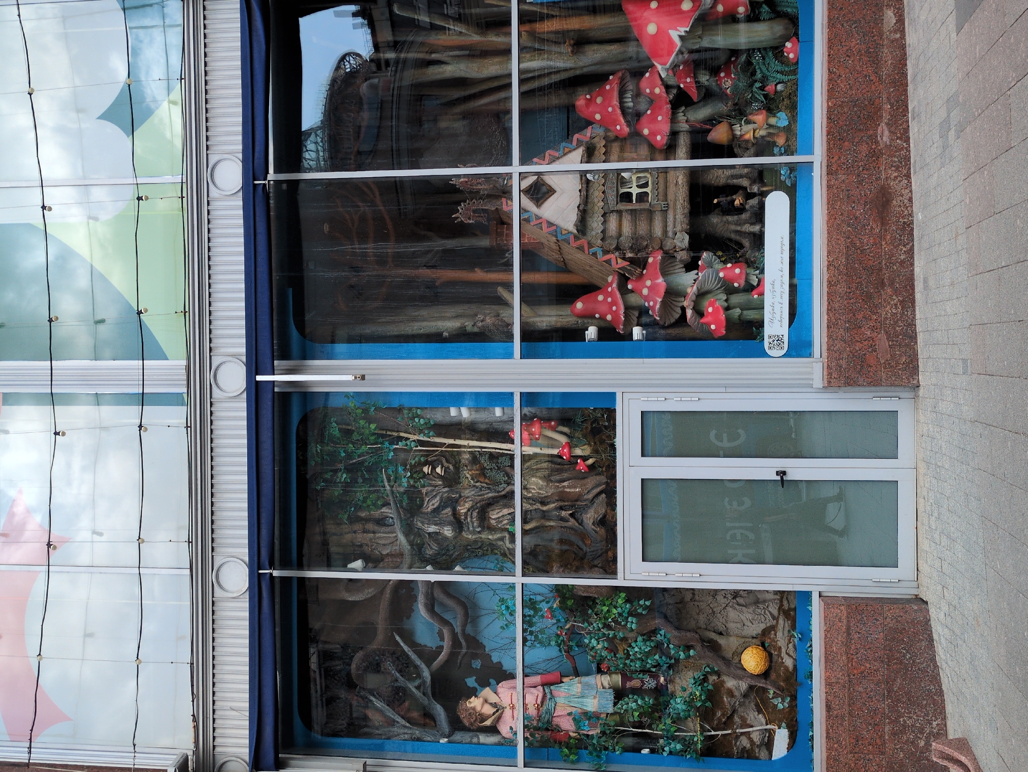 The image shows a storefront display with a whimsical, fantasy-themed scene. The display is divided into two large sections by a vertical mullion.
The left section features a mannequin dressed in a pink and gold outfit, standing amidst greenery and gnarled tree branches. A large, round, golden object is visible on the ground. Behind the mannequin, a large, carved wooden face emerges from a tree trunk, with moss and small red mushrooms around it.
The right section of the display depicts a miniature fairytale village scene. A small, rustic wooden house with a peaked roof and visible windows is at the center. It is surrounded by oversized red and white toadstools, some of which are clustered around the house. Further back, the display shows more tree-like structures and what appear to be other small fantasy elements.
The storefront itself has large glass windows, framed by blue trim. A dark blue awning is visible above the display windows. The building above the display is a multi-story structure with large windows reflecting the sky and surrounding environment. The lower part of the storefront features a base made of reddish-brown, speckled stone. The foreground is a paved walkway.
The image is taken from a low angle, looking up at the storefront. The lighting appears to be natural daylight, with some artificial lights visible on the upper part of the building. There is no visible text that can be clearly deciphered.