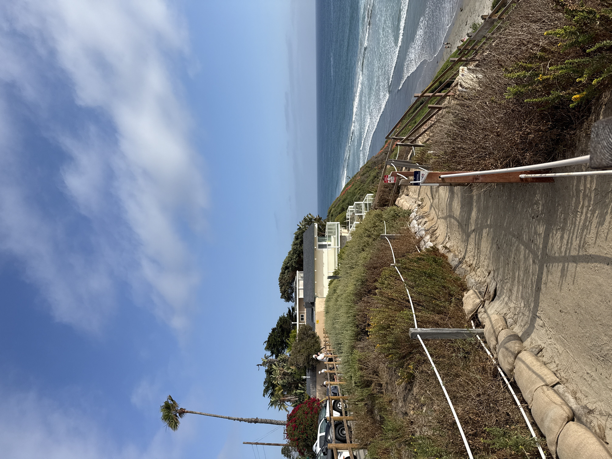 A scenic coastal view in Encinitas, USA, captured on a clear day, showcases a cliffside path, the Pacific Ocean, and coastal properties. In the foreground, a paved or dirt path winds along the top of a cliff, bordered by a simple fence with white ropes strung between wooden posts. Beyond the fence, the cliff drops down, covered in dry, scrubby vegetation, with some greener bushes and visible white sandbags for erosion control.
To the left of the path, further inland, are several multi-story white buildings, characteristic of coastal architecture, surrounded by dense green trees and bushes, including tall palm trees. Cars, including a white SUV, are parked nearby, and a few indistinct figures are visible near the properties.
The vast expanse of the Pacific Ocean dominates the right side of the image, with waves gently breaking onto a sandy beach below the cliff. The ocean fades into a hazy horizon under a bright blue sky dotted with scattered, wispy white clouds.
The clear sky and distinct shadows indicate daytime. Several warning signs are affixed to the fence posts along the cliff path; one clearly reads "DANGER" and "NO TRESPASSING," indicating potential hazards or restricted access. A lone palm tree stands prominently against the sky on the far left, adding to the typical Southern California landscape. The scene evokes a sense of tranquil coastal beauty.