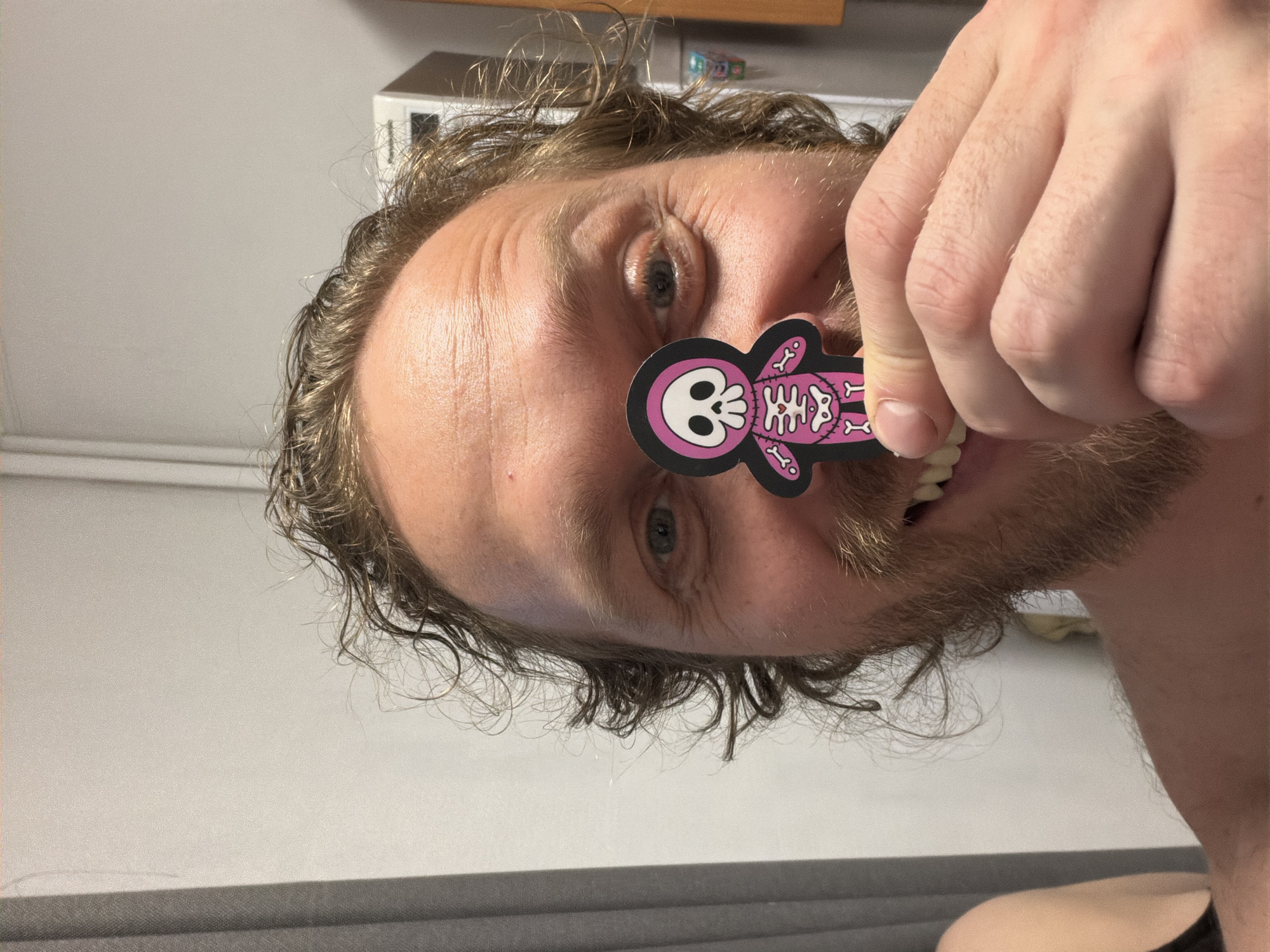 A man with light skin, curly brown hair, and a beard is holding a pink and black skeleton sticker in front of his eye. He is positioned inside a room with a white wall. A piece of furniture is visible in the background. The location of Saint Petersburg, Russia cannot be confirmed from this image.