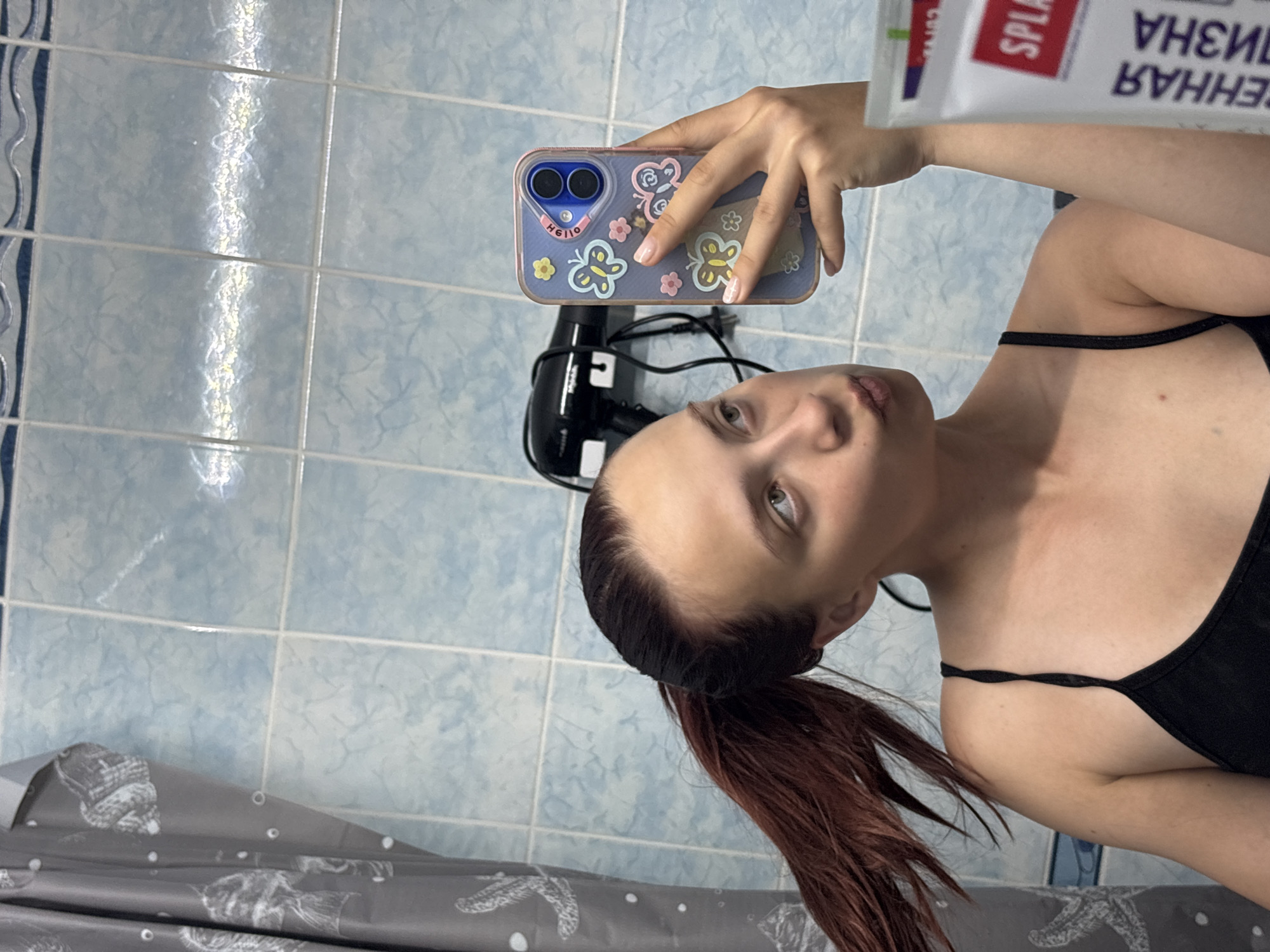 A woman with dark hair and red highlights is taking a selfie in a bathroom. She is wearing a black tank top and holding a phone with a blue case decorated with butterflies. In her other hand, she holds a white tube with the word "SPLAT" on it. A black hair dryer is also visible in the background. The bathroom has blue tiled walls and a grey shower curtain with a fish design. The location may be Прикамский, Russia, based on the text on the product she's holding.