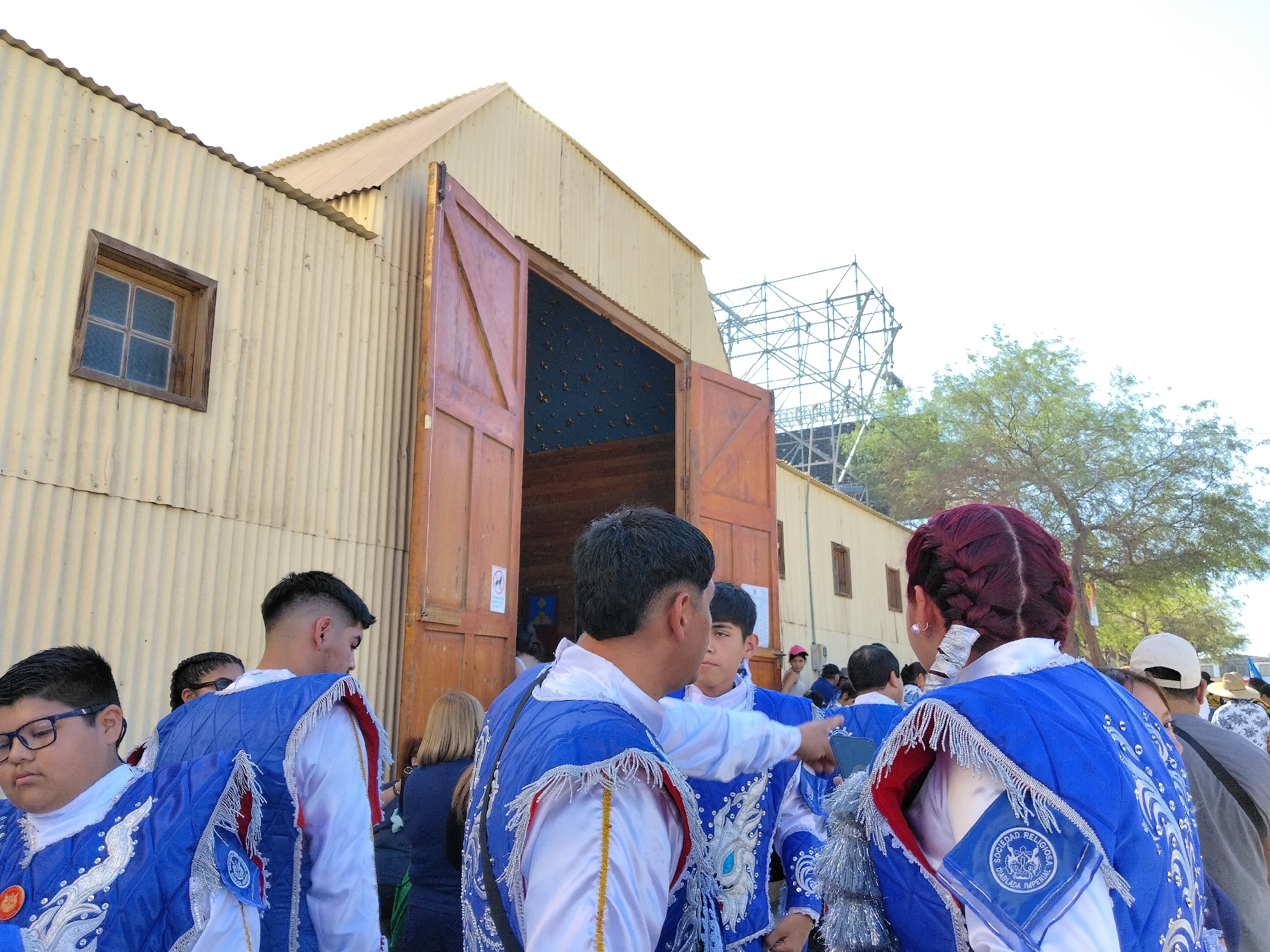 In Pozo Almonte, Chile, a group of individuals in ornate blue and white costumes are gathered outside a corrugated metal building with large wooden double doors. The building has a pitched roof and a small window. Some of the individuals are wearing these decorative outfits, which feature elaborate embroidery and silver fringe. One individual, facing away from the camera, points towards the right. The sky is bright and overcast, and a metal scaffolding structure is visible in the background behind a tree. The scene appears to be a public gathering or event.