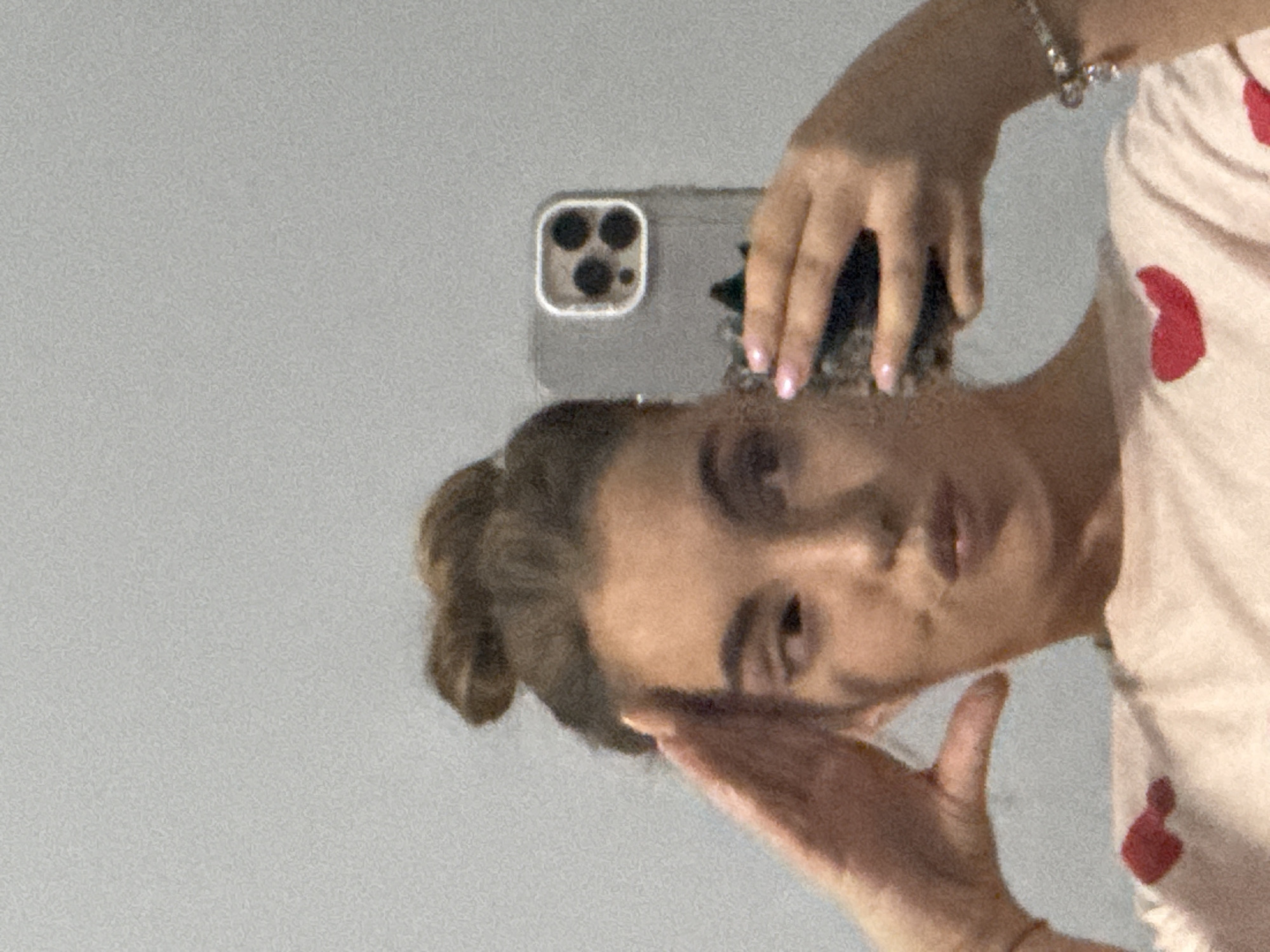 A candid mirror selfie depicts a young female from the chest up, positioned against a plain, light grey wall. Her brown hair is styled in a high, messy bun. She is wearing a light-colored t-shirt with multiple red heart patterns. Her left hand is raised, with fingers splayed and palm resting against her temple, while her right hand holds a silver-colored smartphone encased in a clear cover, revealing a triple-lens camera system on its back. Her fingernails are painted light pink. A beaded bracelet with charms is visible on her right wrist. Her gaze is directed slightly to her right, and her lips are softly puckered. The image exhibits noticeable digital grain. The scene is situated in Sofia, Bulgaria, as specified.