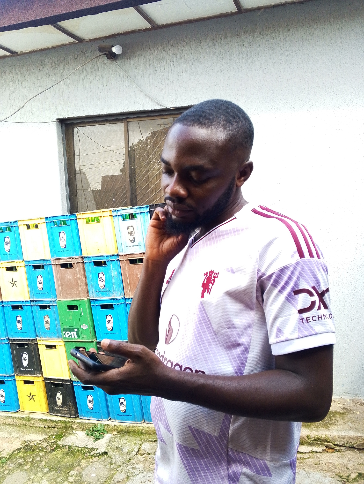 An outdoor medium shot shows a Black man in his late 20s or early 30s standing in front of a stack of colorful plastic crates and a textured white wall. The man, with a short haircut and a beard, is looking down at a smartphone he holds in his right hand, with his left hand touching his ear. He is wearing a white jersey with purple stripes on the sleeves and a purple logo on the left chest, and a graphic design on the lower part of the sleeves. The crates are stacked in an irregular pattern, with rows of blue, yellow, brown, and green crates. A window with blinds is visible on the wall behind him, and a bare lightbulb is mounted on the wall above the window. The ground in front of the crates is covered with a mix of dirt, moss, and small stones.