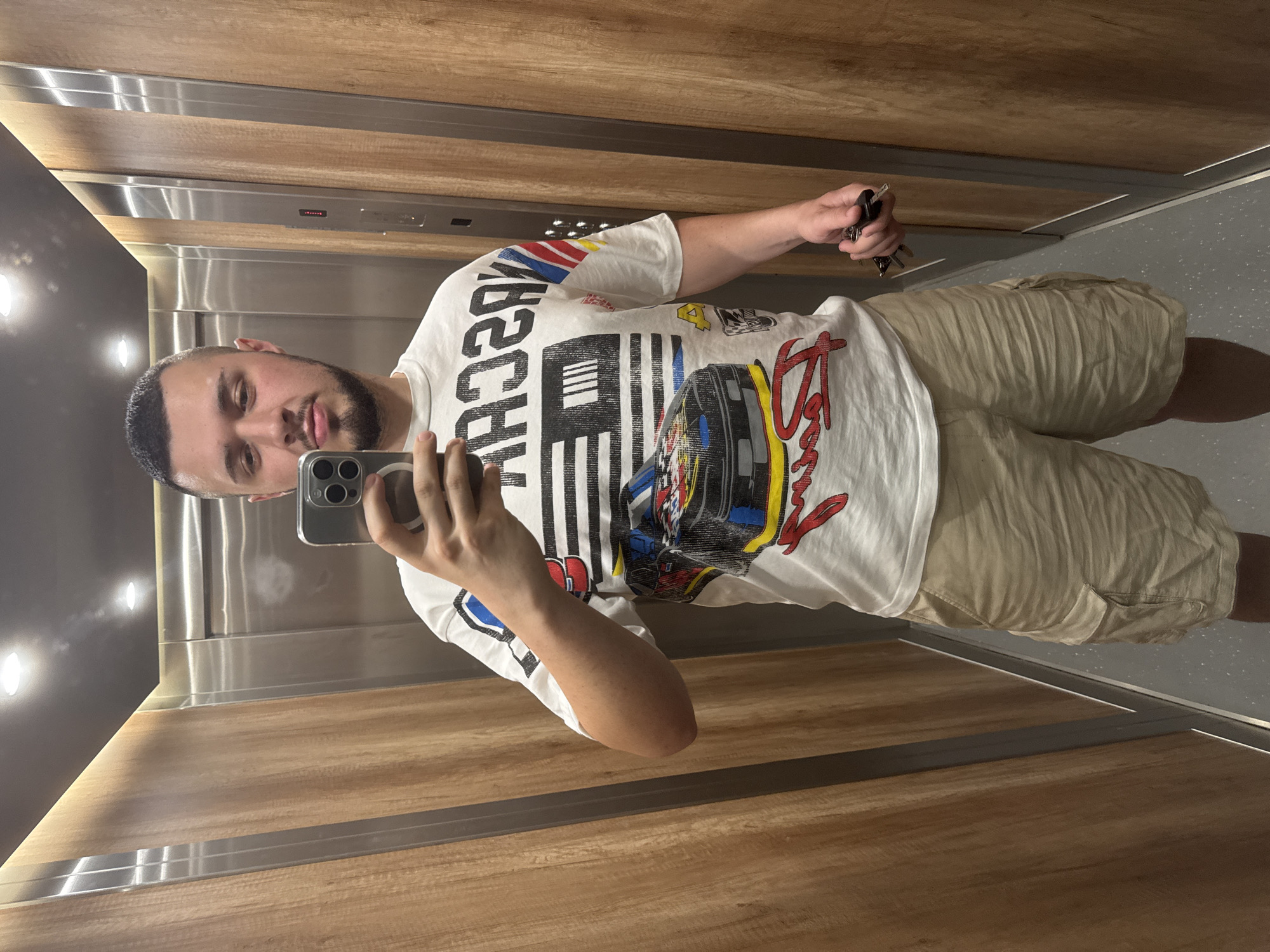 A young man with short dark hair and a beard stands in an elevator, taking a mirror selfie with his phone. He is wearing a white t-shirt with a graphic print featuring a race car and the word "NASCAR," and khaki cargo shorts. In his right hand, he holds a set of keys. The elevator walls are paneled with light-colored wood and have stainless steel accents. The floor is grey with a speckled pattern. The lighting in the elevator is from overhead fixtures.