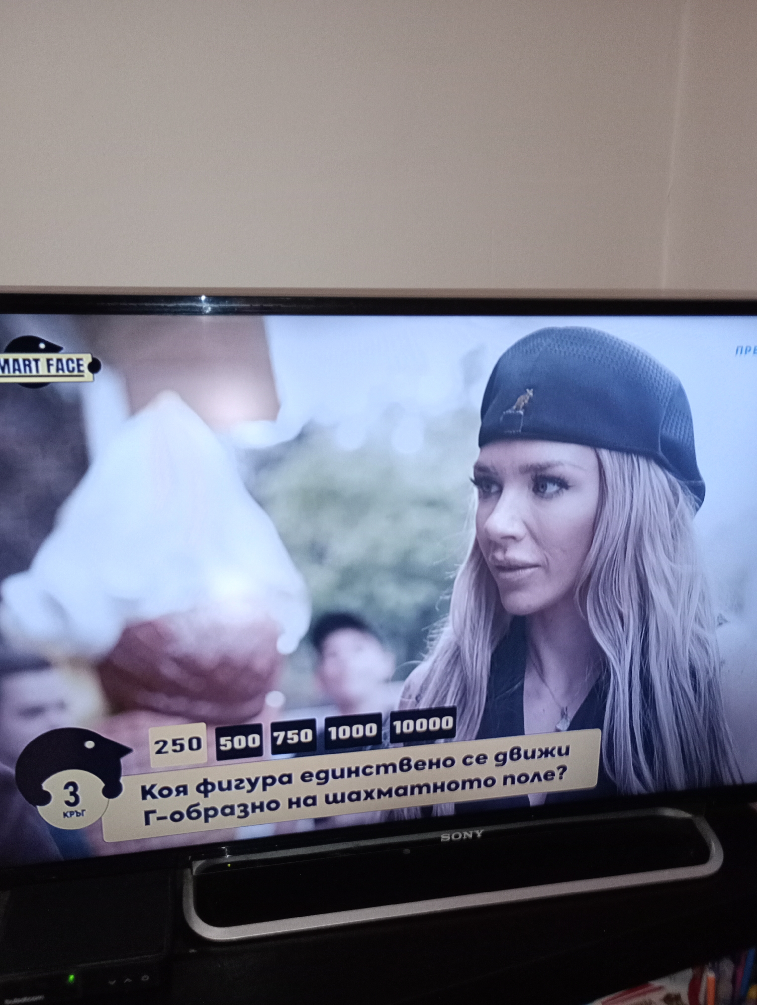 The image is a television screen displaying a game show or quiz program. On the left side of the screen, a logo with the text "SMART FACE" is visible. Below this, a graphic of a black bird with the number "3" and the Cyrillic word "кръг" (meaning "round") is displayed. To the right of this graphic, there are rectangular boxes with numbers: "250", "500", "750", "1000", and "10000".
Below the numbers, a question is displayed in Cyrillic text, which translates to "Which figure moves exclusively in a G-shape on the chessboard?". This indicates a question related to chess.
The right side of the screen shows a close-up of a woman with blonde hair, wearing a dark beret. She is looking to her left with a neutral expression. The background on the right appears to be blurred greenery.
The bottom of the television screen shows the "SONY" logo. The image is a photograph taken of the television screen, likely in a home environment, given the surrounding wall and what appears to be part of a television stand or furniture. There is no indication of the city of Harmanli, Bulgaria in the image.