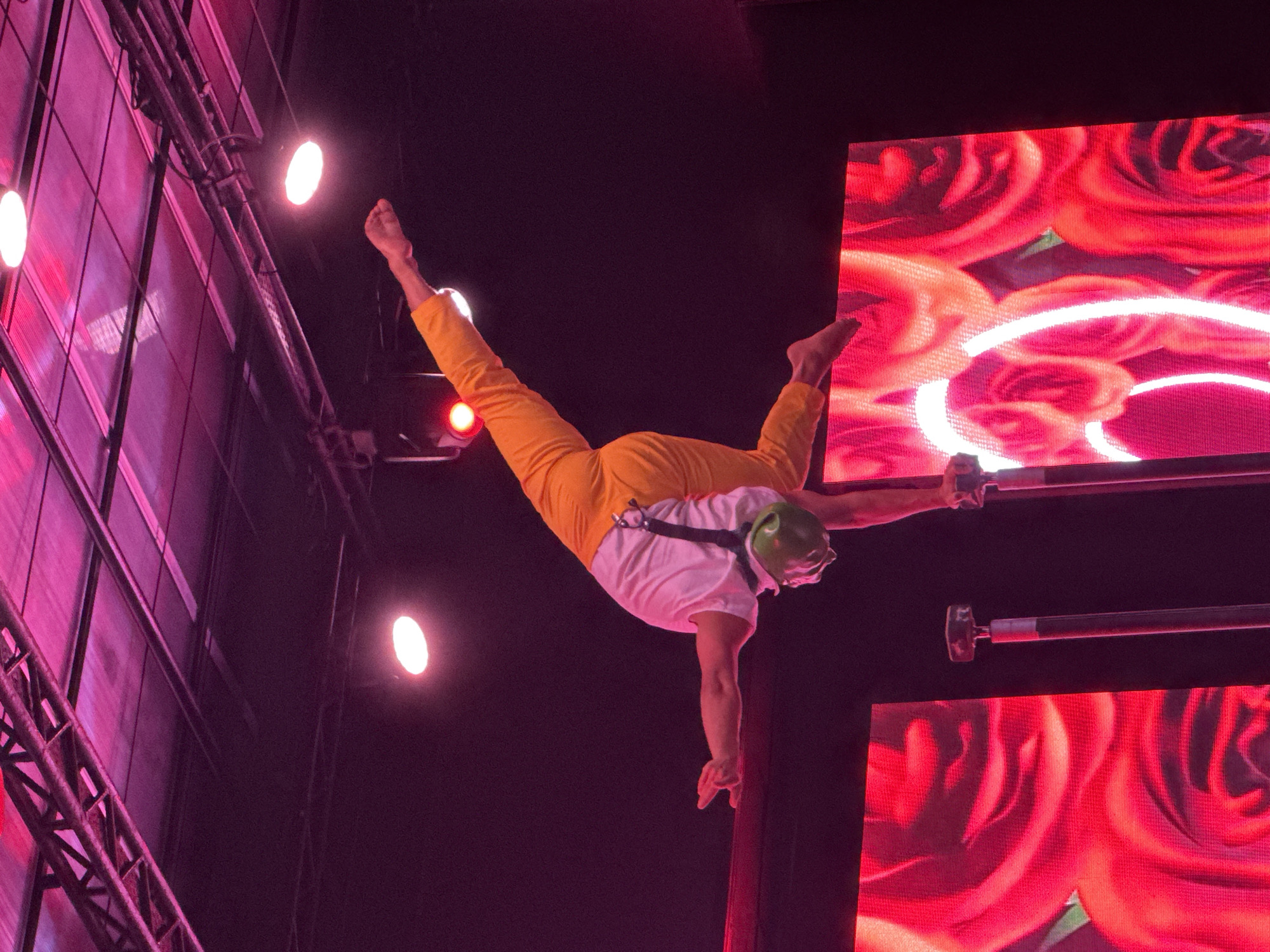 A lone male acrobat in a white t-shirt and orange pants performs a handstand on two vertical poles during a nighttime performance. He is wearing a green helmet. His body is extended, with his right arm reaching upwards and his left arm outstretched to the side, fingers splayed. His legs are in a V-shape, with his right leg extended higher than his left. In the background, there are large screens displaying images of red roses, illuminated by pink and purple stage lighting. The ceiling of the venue is visible, with exposed metal beams and track lighting. The overall atmosphere is that of a live performance. The location is the town of Manavgat, Turkey, where a circus event is taking place.