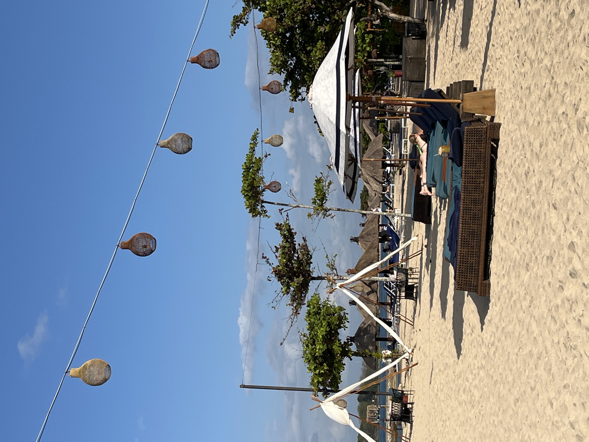 The image depicts a serene beach scene in Kuta Selatan, Indonesia. The foreground is dominated by a sandy beach with several lounge chairs and umbrellas, suggesting a resort or beach club setting. A person is reclining on one of the lounge chairs, facing away from the camera, appearing relaxed. In the mid-ground, more lounge chairs, thatched-roof umbrellas, and small tents are visible, indicating a shaded area for patrons. Trees with lush green foliage are scattered across the beach, providing natural shade. Overhead, a string of decorative gourd-like lanterns hangs against a clear blue sky. The background shows a body of water, likely the ocean, and a distant shoreline with more vegetation. The overall atmosphere is one of tranquility and leisure, typical of a tropical beach destination.