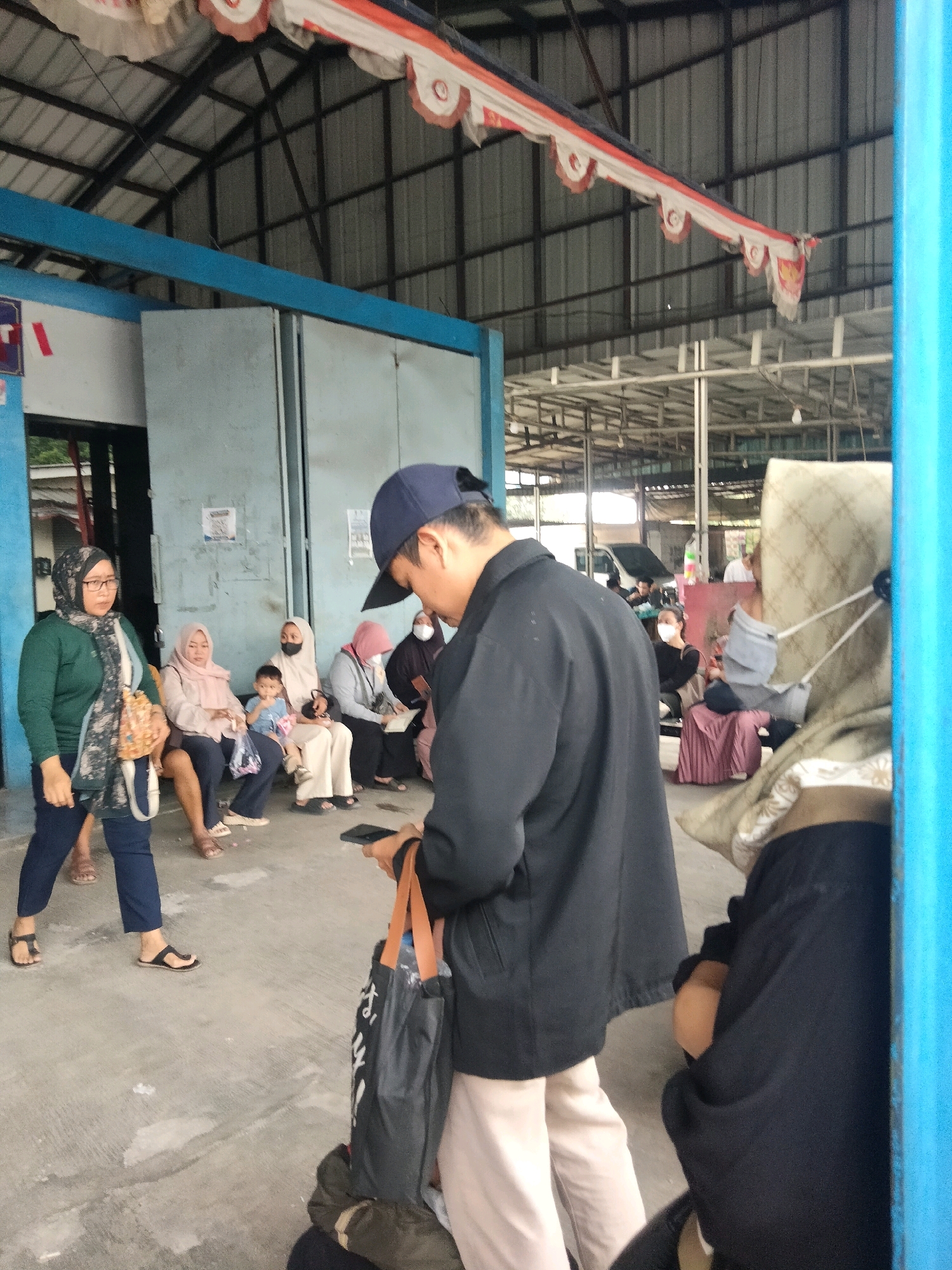 The image shows a group of people waiting at what appears to be a terminal or bus station in Sukaasih, Indonesia. In the foreground, a person wearing a black jacket and a blue baseball cap is holding a black bag and a mobile phone. They are standing on a concrete floor in front of a blue structure. To the left, a woman in a green cardigan and headscarf is walking. Several other individuals, including women and children, are seated on benches or standing in the background. The environment is an open-air covered area with a metal roof supported by beams. Decorative banners are hung from the ceiling. The overall scene depicts a moment of waiting or transit.