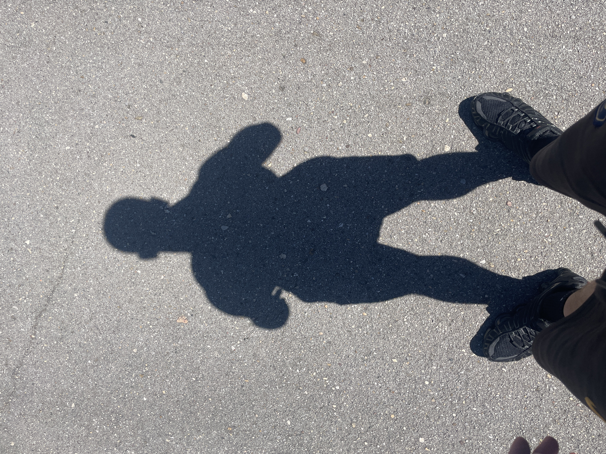 A high-angle shot shows a shadow of a person on asphalt. The person is wearing shorts and shoes, which are also visible in the frame. The person is likely standing and holding a phone or camera to take the photo. The location appears to be an outdoor area with asphalt pavement. There are no signs to determine location in Sofia, Bulgaria.