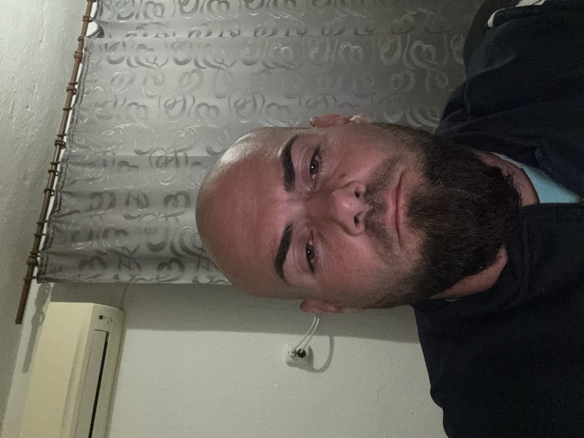 A bald man with a beard is facing the camera indoors. He is wearing a dark-colored jacket over a light-colored shirt. Behind him is a wall with a patterned window shade. An air conditioning unit is visible on the wall to the left. There are cords hanging from the wall near the man's ear. There is no indication of the location or interaction of the individuals.