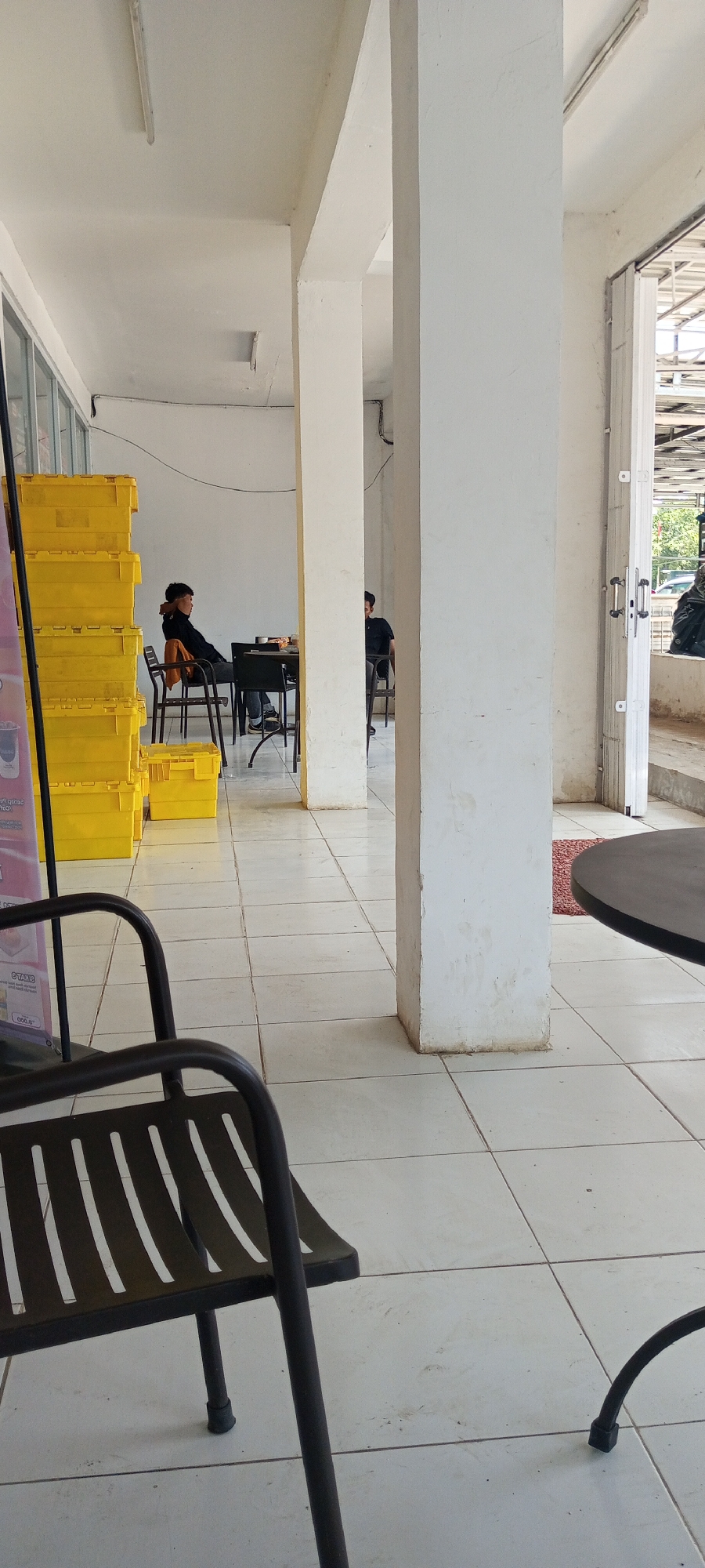 The image depicts an outdoor seating area with tiled flooring, likely located in Lempa, Indonesia, given the context. The scene is dominated by white pillars and a white ceiling with fluorescent lighting fixtures. In the background, several stacks of bright yellow storage containers are visible on the left side.
Two individuals are seated at tables in the middle ground. The person on the left is a young male wearing a dark shirt and jeans, seated at a table with a dark chair. He appears to be looking down. To his right, another male, also in a dark shirt, is seated at a table further back.
In the foreground, a dark metal chair with a slatted seat and back is positioned prominently, facing towards the left of the frame. Another dark metal chair leg is visible on the far right, suggesting another table or seating arrangement.
The environment appears to be an open-air structure, with a large doorway or opening on the right side revealing a glimpse of the outdoors, including a grey corrugated roof and some greenery. A small red rug is partially visible on the floor near the pillars. The overall impression is of a casual, functional outdoor space.