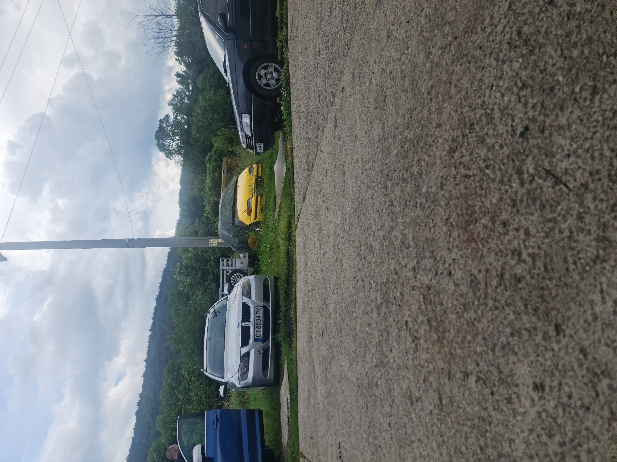 The image, best viewed rotated 90 degrees counter-clockwise from its original orientation, captures an outdoor scene in Buzovgrad, Bulgaria, featuring several vehicles parked alongside a concrete or asphalt surface.
In the foreground, a silver BMW, possibly a 5-series sedan (E60 generation), is parked on a grassy verge. Its front license plate, "CT 8034 PX," is clearly visible. Behind the BMW, the open door of a blue car reveals a person partially visible inside, suggesting someone is entering or exiting the vehicle. Further along the grassy verge, a dark-colored sedan, resembling an older model, is parked. Behind it, a bright yellow car is partially covered by a dark tarp. A white trailer with a spare tire mounted on its side is also visible in the mid-ground. A tall utility pole stands to the left of the scene.
The background consists of dense green trees and a forested hill or mountain range, stretching across the horizon. Above the trees, the sky is overcast with fluffy white and grey clouds, indicating daylight hours with soft, diffused light. The overall setting appears to be a rural or semi-rural area, possibly a private drive or a less-trafficked road, with vehicles parked off the main thoroughfare amidst natural surroundings.