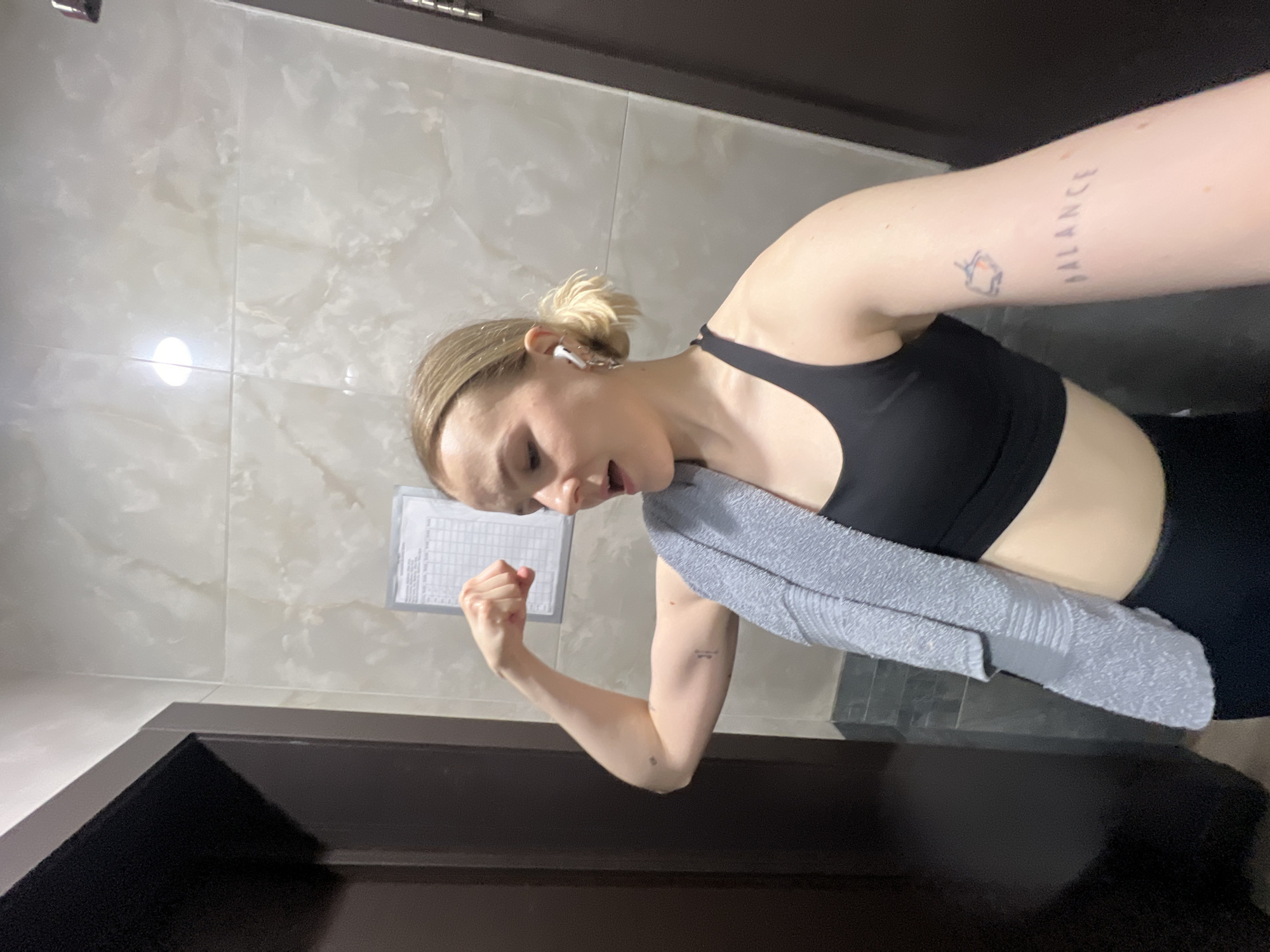 A fair-skinned woman in athletic attire stands in what appears to be a bathroom with light-colored tile walls. She wears a black sports bra and leggings, with a gray towel draped around her neck. An earpiece is visible in her ear. She flexes her left bicep, revealing a tattoo on her left forearm. Another tattoo is present on her right forearm, displaying the word "BALANCE" along with a small image. A chart or schedule is affixed to the wall behind her. The lower portion of a dark-colored door frame is visible in the foreground. The location is in the city of Khimki, Russia.