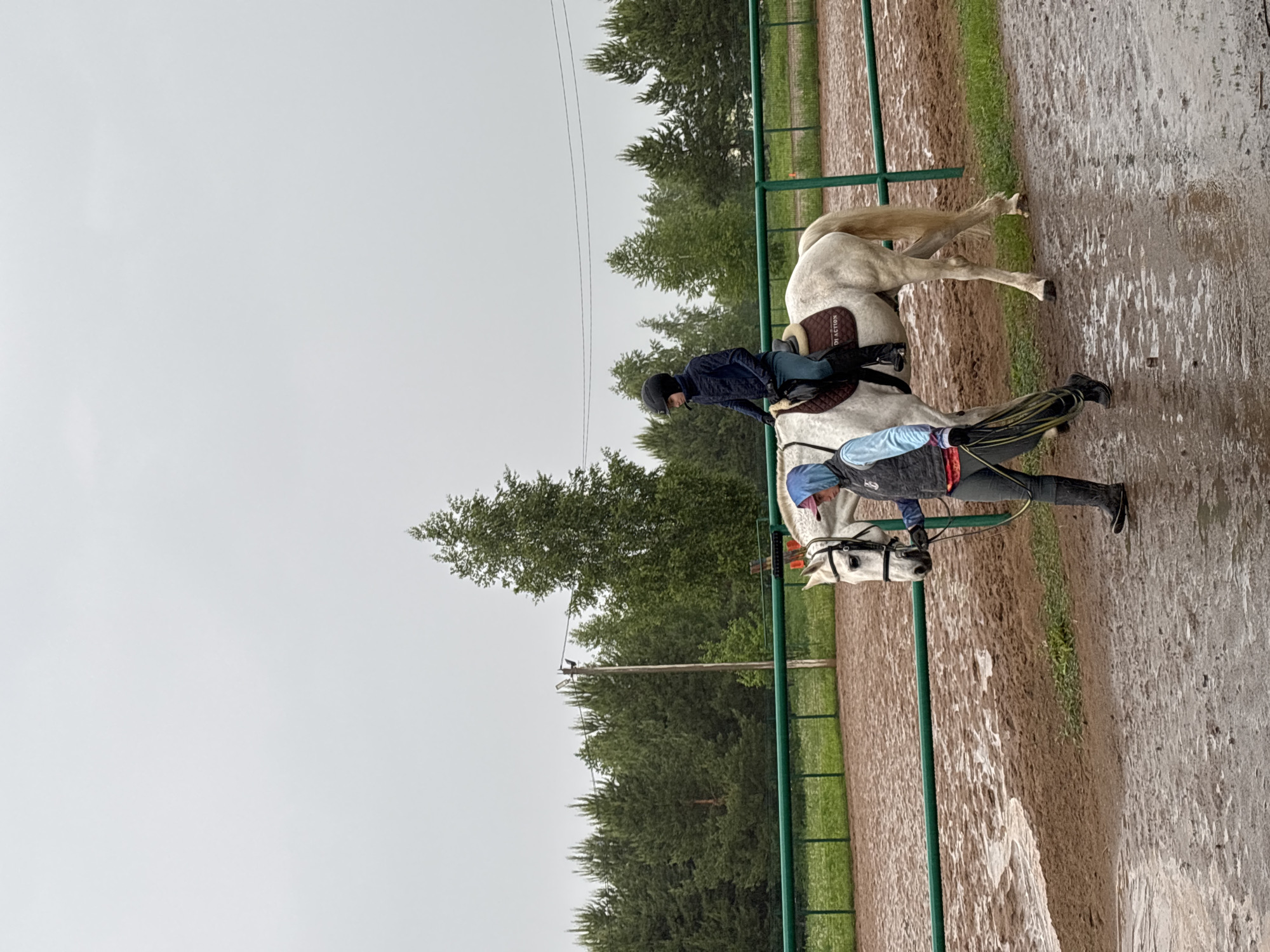 The image shows two people with a white horse in what appears to be a riding arena or training area. A person wearing a helmet and dark jacket is on the back of the horse, while another person wearing a blue hat and a vest is standing near the horse's head, possibly leading it. The arena is bordered by a green fence. The ground is muddy. Trees are visible in the background, and the sky is overcast. The image was taken in the city of Русский Вожой, Russia.
