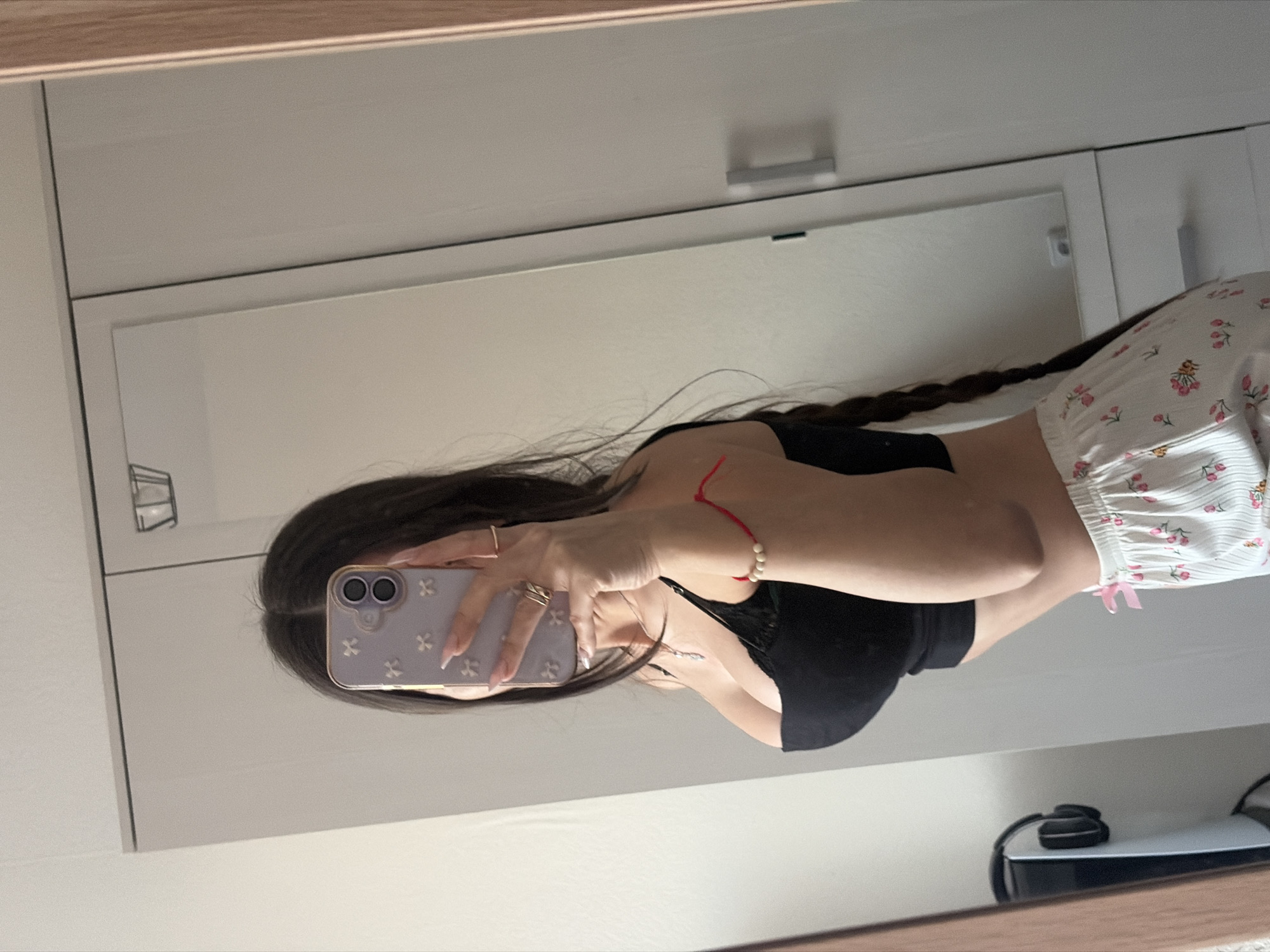A female is reflected in a mirror, presumably taking a selfie with a lavender-colored phone case adorned with a floral pattern. She has dark hair in a braid, wearing a black top and white shorts with a floral design. Her left hand has multiple rings. A red string bracelet is on her right wrist. In the background, there's a white cabinet and a black PlayStation 5 on a white surface. There is no indication that this photo was taken in Saint Petersburg, Russia.