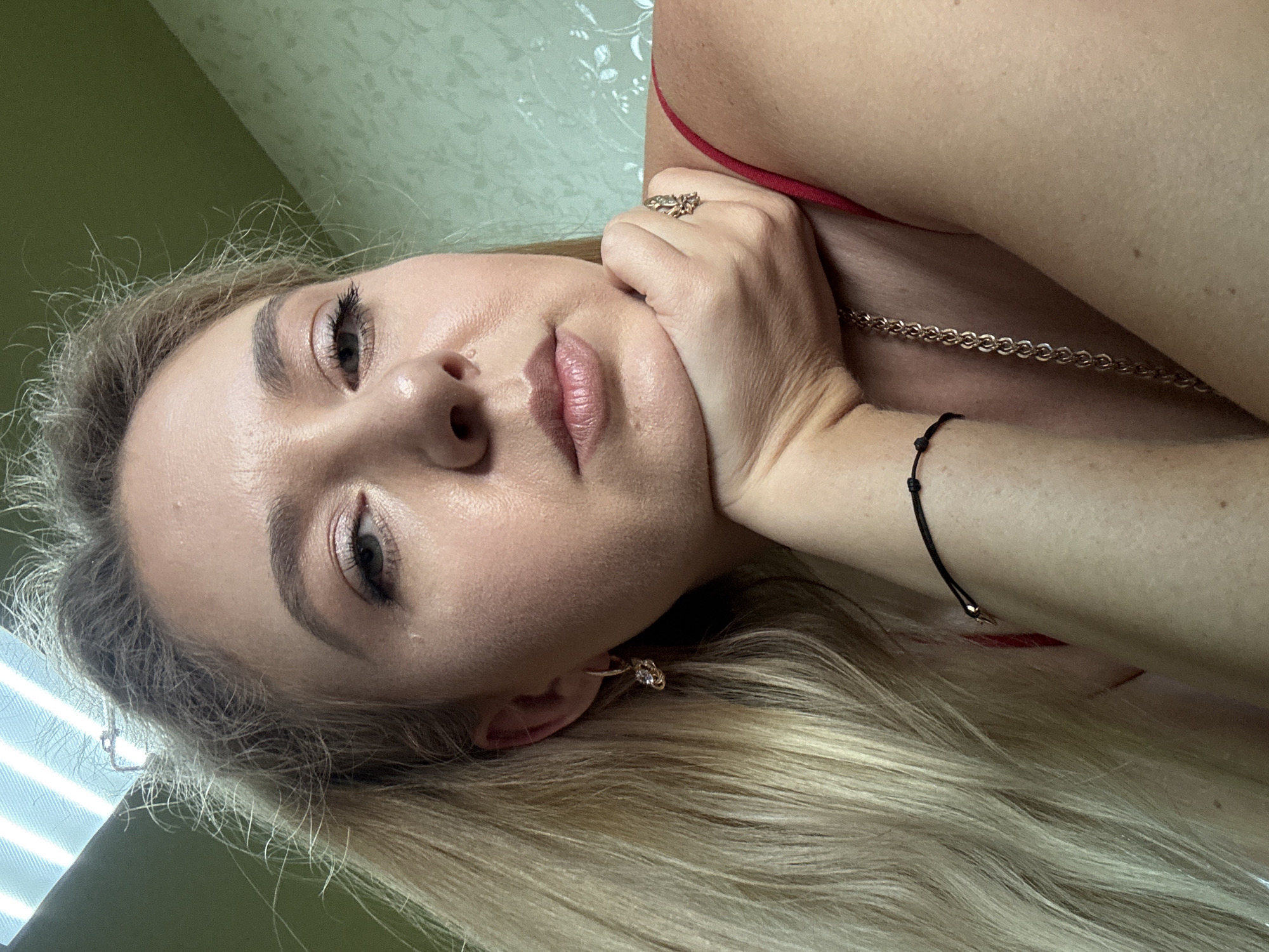 A close-up, selfie-style portrait shows a blonde woman with her right hand supporting her chin. She is wearing a red top, a gold chain necklace, a thin black bracelet with beads, a gold earring with a diamond, and a gold ring on her right hand. The background is a light green wall with a floral pattern and a ceiling with fluorescent lighting. The woman's hair is long and blonde, and it falls over her left shoulder. Her eyes are open and looking at the camera. She has makeup on her face, including eyeshadow and lipstick.
