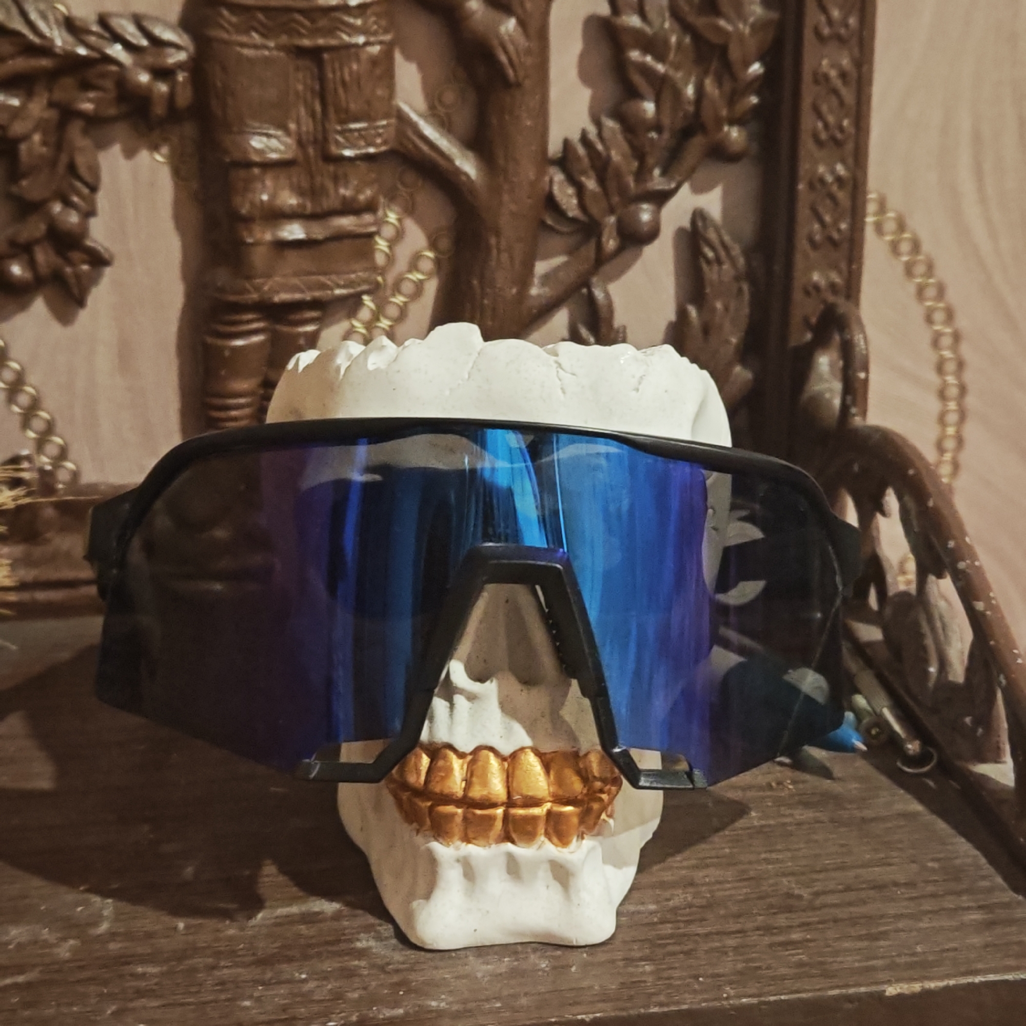 A still-life photograph shows a white skull with gold teeth wearing black sunglasses with reflective blue lenses. The skull rests on a dark wooden surface. In the background is a detailed, brown-toned wooden sculpture or carving. A gold chain detail appears behind the skull, near the top of the sculpture. The sculpture depicts figures. The location, Kladovitsy, Russia, is irrelevant to the visual content of the image.