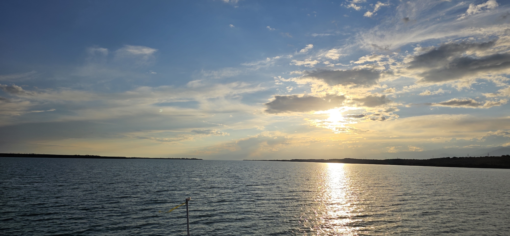 The image depicts a wide expanse of calm water, likely a lake or reservoir, with a low, tree-lined horizon in the distance. The sky is a dominant feature, filled with scattered clouds that are illuminated by the setting sun. The sun itself is partially obscured by clouds, casting a golden glow and creating a shimmering reflection on the water's surface. The overall scene conveys a peaceful and serene atmosphere. It is plausible that this scene is located near Pristan'-Przheval'sk, Kyrgyzstan, given the natural landscape and potential for such bodies of water in the region. There are no people or specific actions visible in the image; the focus is on the natural environment.