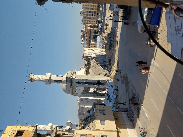 The image displays an urban landscape in Khan Yunis, Palestinian Territory, characterized by extensive structural damage under a clear blue sky. A large, light-colored mosque with a prominent central dome and a tall, multi-tiered minaret stands in the mid-ground, exhibiting significant damage, including collapsed sections of its walls and surrounding structures, with rubble accumulated at its base. A second, shorter minaret is partially visible to the left of the main mosque.
Numerous multi-story buildings frame the scene, many showing severe damage with partial collapses, exposed rebar, and large piles of concrete and debris at ground level. Some buildings in the background appear relatively intact.
The ground consists of paved roads with faded markings, intermingled with substantial areas of rubble and debris. Approximately a dozen individuals are visible walking across the scene. In the foreground, a group of five individuals, including adults and children, walks on the paved road. Other individuals are seen walking further into the scene, navigating areas with debris. On the left side of the foreground, a bicycle is visible near a partially damaged building. On the right, temporary structures with blue tarpaulins are present. A large white tent structure is located in the mid-ground among the damaged buildings. A single dark wire stretches horizontally across the upper portion of the frame. Shadows indicate clear sunlight.