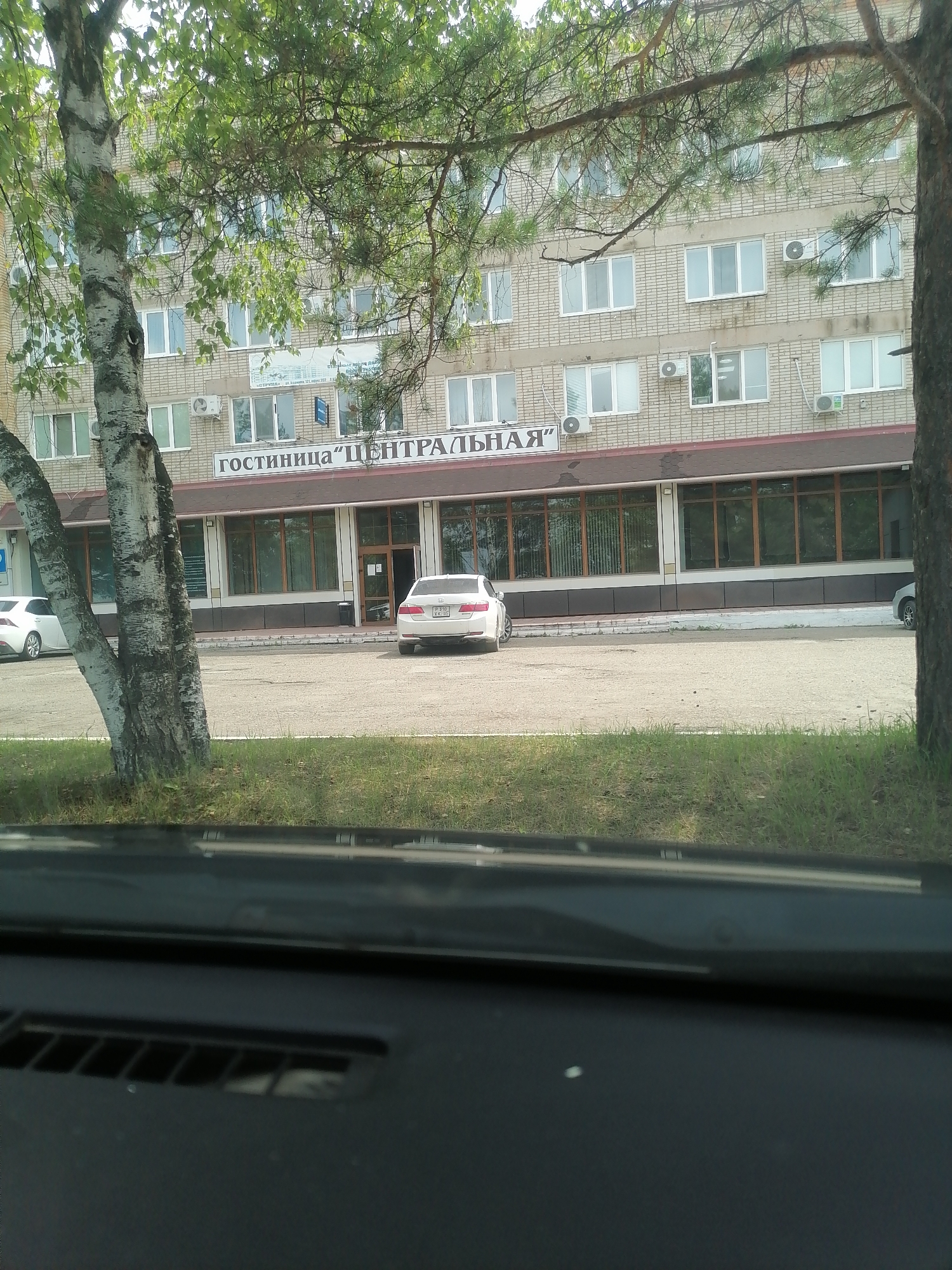 This is an eye-level outdoor shot, taken from inside a vehicle, of a three-story brick building that houses the "Centralnaya" hotel in Lesozavodsk, Russia. The building features numerous windows and air conditioning units. In front of the hotel, a white sedan is parked, and a white hatchback is parked to the left. Trees are visible in the foreground, obscuring parts of the building. The sky is overcast.