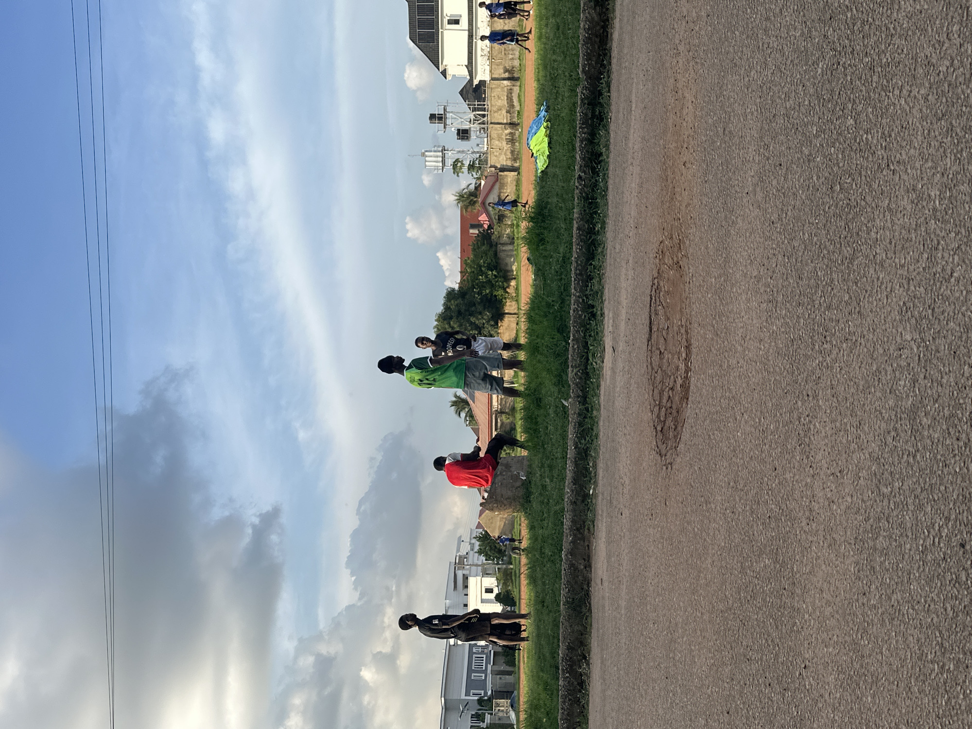 The image shows a group of young men gathered outdoors in a residential area of Abuja, Nigeria. In the foreground, a damaged asphalt road is visible with a large pothole. Beyond the road, a grassy verge leads to a field where several individuals are present. Some are standing and observing, while others appear to be engaged in or preparing for an activity. One person is seated on a rock. In the background, modern residential buildings and water towers suggest an urban or suburban setting. The sky is partly cloudy, indicating daytime. The overall impression is a casual gathering in a neighborhood.