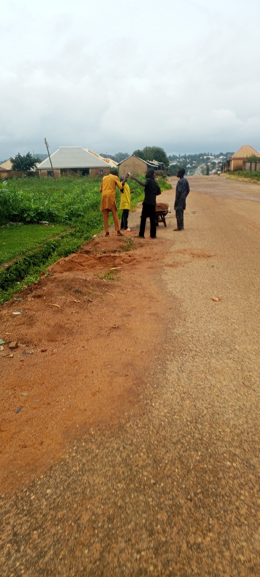 In Bahit, Nigeria, four individuals stand on a dusty, unpaved road, partially obscured by motion blur. A man in a yellow traditional outfit faces away from the camera, his right arm extended. To his left, a person in a yellow garment stands, and another, dressed in black, gestures with their right hand. To the far right, a fourth person in a dark, patterned traditional outfit stands facing left. They are positioned near a low wooden table laden with a brown substance, possibly soil or sand. The road is bordered by a patch of green vegetation on the left and houses in the background under a cloudy sky. The ground is a mix of reddish-brown soil and the rough texture of the road.