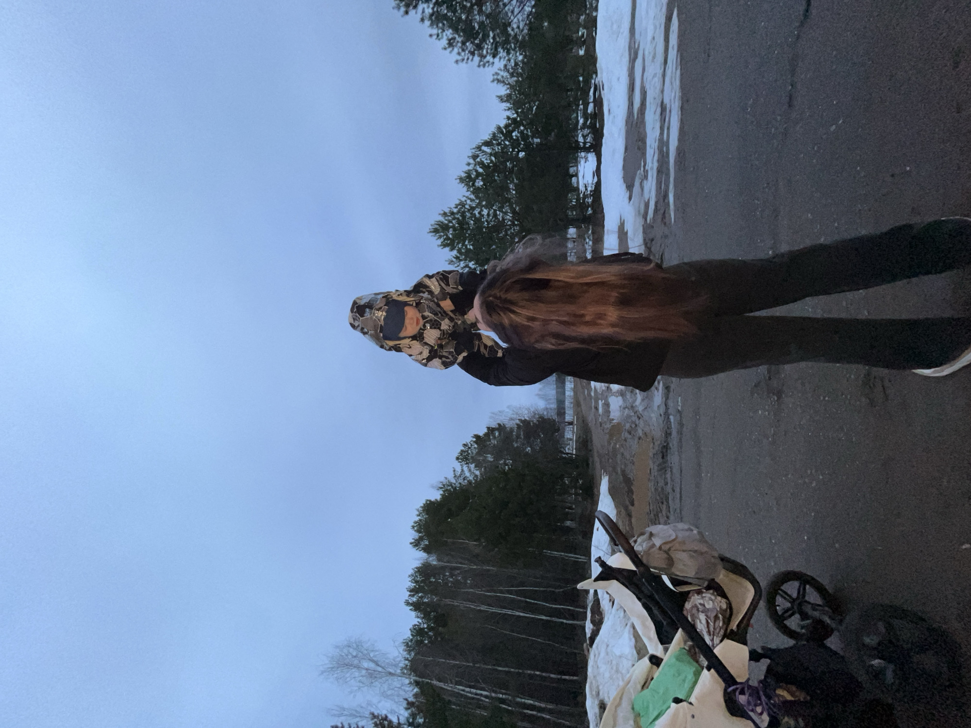A medium shot depicts an adult holding a child outdoors. The adult is wearing a patterned jacket and dark pants. The child is partially obscured by the adult.
The location appears to be a roadside in a wintry environment. Patches of snow are visible. Trees are in the background. A stroller is present in the foreground, containing items covered by fabric.
The city of Chaikovsky, Russia is not visually confirmed in the image.