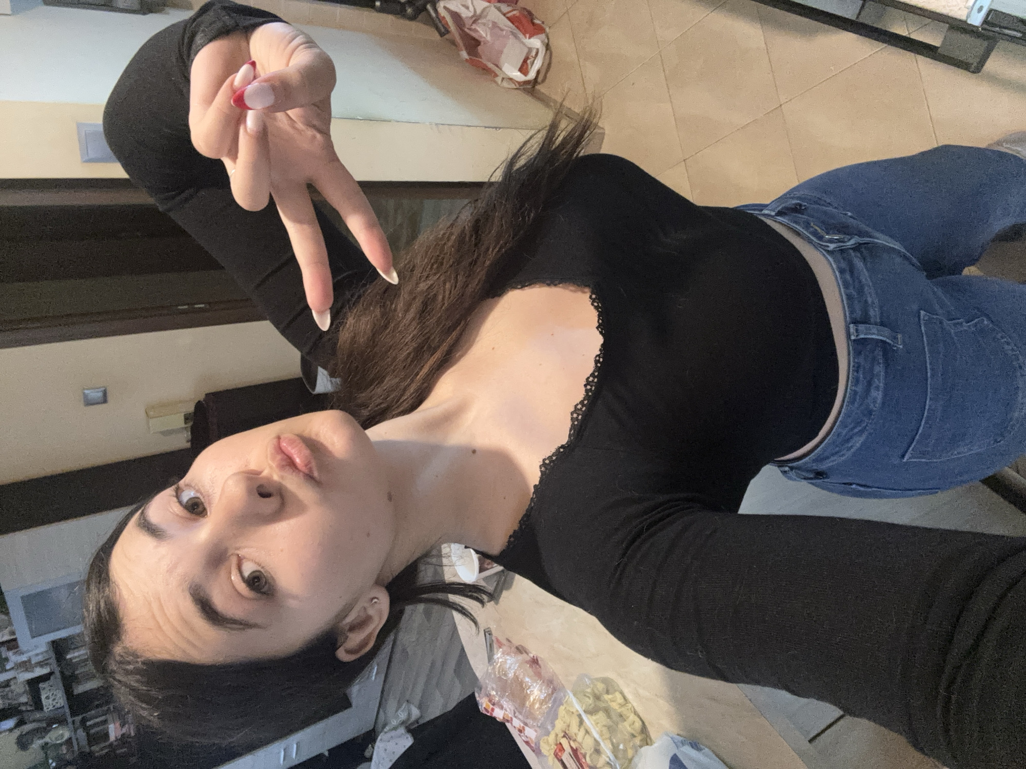 A young woman is captured in a selfie taken indoors, likely in a home setting in Plovdiv, Bulgaria. She is positioned with her head tilted, looking directly at the camera with a slight pout on her lips, conveying a casual and playful mood. Her long, dark hair falls over her shoulders.
She is dressed in a black long-sleeved top featuring a delicate black lace trim along the neckline, paired with blue denim jeans. Her right hand is raised, forming a "peace" sign with her index and middle fingers. Her nails are long, styled with a French manicure, characterized by white tips and a distinct red accent on the very ends of the tips. A small silver stud earring is visible in her left ear.
The background of the image reveals a light-colored tiled floor. Other elements include a glimpse of what appears to be a table or counter with a clear plastic bag containing light-colored items, possibly snacks or pasta, and a wall featuring a light switch or electrical outlet. The ambient lighting suggests the photo was taken during the daytime. No visible text is legible within the image.