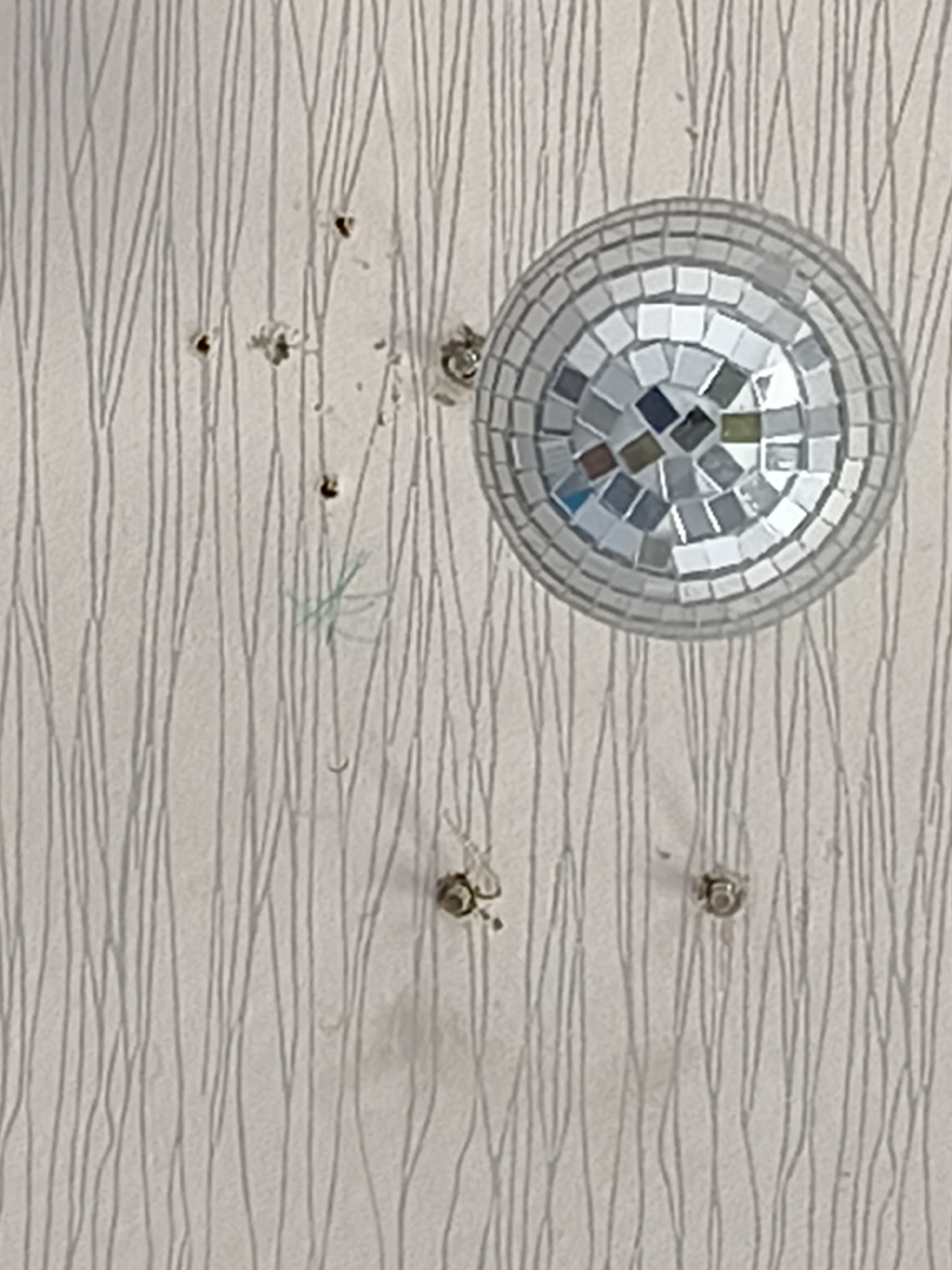 The image shows a close-up of a wall with a textured, light-colored wallpaper featuring thin, vertical grey lines. A small disco ball, composed of numerous square mirrored tiles, is attached to the wall. Several small, dark circular marks or holes are scattered across the wall, some with debris around them, suggesting they may be from a projectile. In the lower right quadrant of the image, a small circular metal object, possibly a screw head or a fastener, is visible. There are no people or discernible Moscow landmarks present in the image.