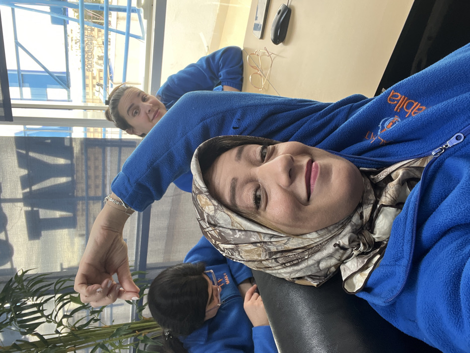 Three women are depicted in an indoor professional environment in Meknes, Morocco.
The foreground subject, a woman wearing a multi-toned patterned headscarf and a blue fleece zip-up jacket with an "Abila" logo embroidered on the right chest, smiles directly at the camera from a black chair. To her right, a second woman in a similar blue fleece jacket looks towards the camera with a subtle smile, her right arm raised, with a silver bracelet visible on her wrist, and her hand making a downward gesture. She is seated at a light-colored table, which holds gold-framed eyeglasses, a black smartphone, and a black computer mouse. To the left, a third woman, also in a blue fleece jacket and wearing glasses, is seated and focused downwards at the table.
Through large background windows, the exterior of a building with the word "POLYCLINIQUE" visibly printed on it is clearly discernible, establishing the immediate vicinity as a polyclinic in Meknes. Blue architectural elements and a distant urban landscape are visible outside the windows. An indoor potted plant stands on the far left of the frame, and the shadow of large letters, possibly "TAYA," is cast onto an internal window blind.