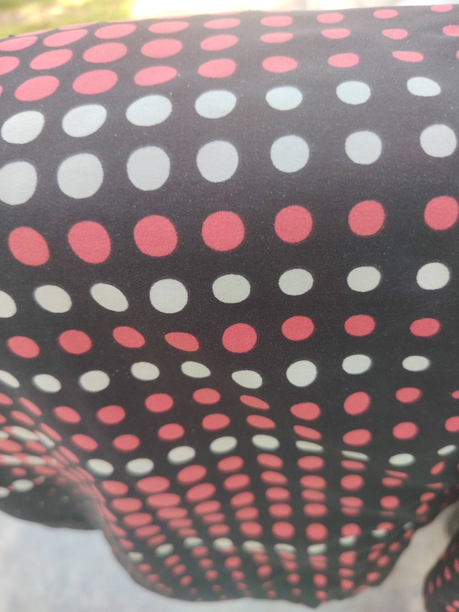 A close-up shot captures a dark brown or black fabric adorned with a polka dot pattern. The dots are arranged in alternating rows of light pink/coral and off-white/light gray, set against the dark background. The fabric is draped and curved, creating a three-dimensional effect that distorts the circular shape of some dots, especially towards the bottom and sides of the frame.
The top portion of the image reveals a highly blurred background of green and yellow hues, indicative of foliage or grass in an outdoor setting. The background at the bottom is similarly out of focus, appearing lighter, potentially suggesting another distant outdoor element. Natural daylight illuminates the fabric, casting a subtle sheen on the dots. The texture of the fabric shows slight creasing and minor imperfections, such as small lint particles. No people, specific activities, or discernible text are visible in the image.