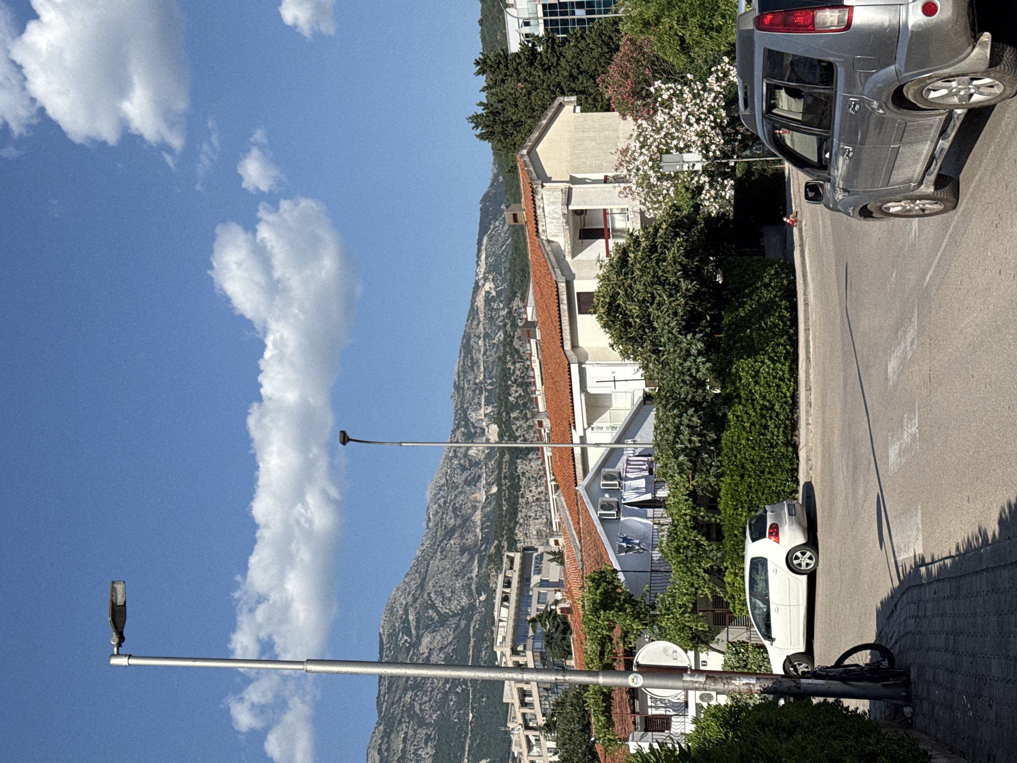 This is a vertical, eye-level shot of a street scene in Budva, Montenegro, under a bright blue sky with scattered white clouds. The sun casts sharp shadows, indicating it's daytime. A grey SUV is parked on the right side of the asphalt road. Further down the road, a white hatchback is also parked. A bicycle is leaning against a lamppost in the foreground.
The scene includes buildings with red-tiled roofs, some with balconies, and lush green trees and bushes. In the background, a rugged, mountainous landscape rises steeply. The architecture suggests a Mediterranean coastal town. There are no people clearly visible in the frame. The overall atmosphere is one of a quiet, sunny day in a scenic, populated area.