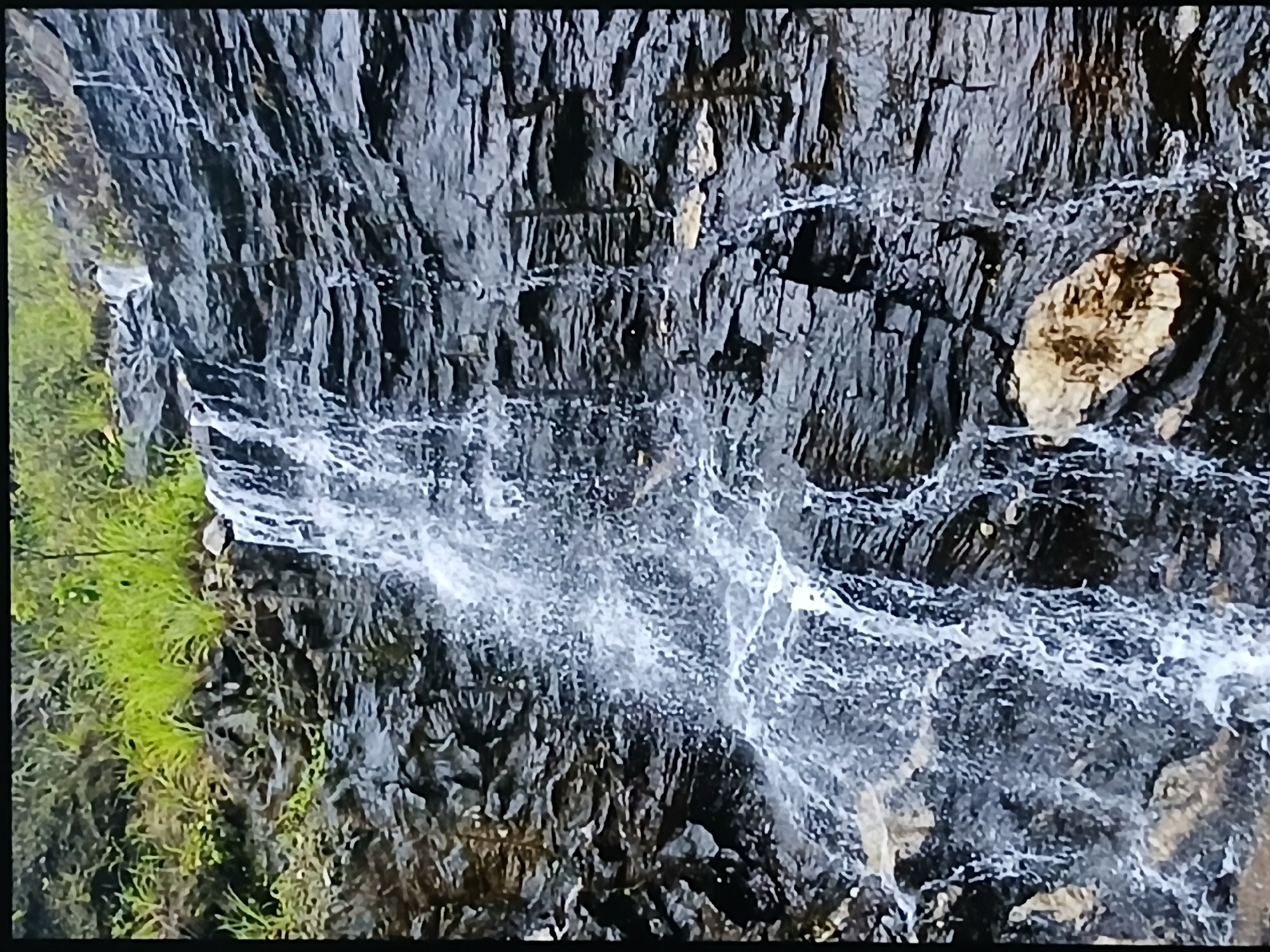 The image depicts a waterfall cascading down a dark, rocky cliff face, with green vegetation clinging to the rocks on the left. The water appears turbulent, creating a white, frothy appearance. The rock face is primarily dark grey to black, with some patches of lighter, possibly yellowish-brown, rock. The scene appears to be outdoors, possibly in a natural environment.
While the image doesn't explicitly provide information to confirm that it is from the city of Bengaluru, India, no definitive evidence contradicting this is present in the image. Given the prompt's assertion, the location can be assumed.