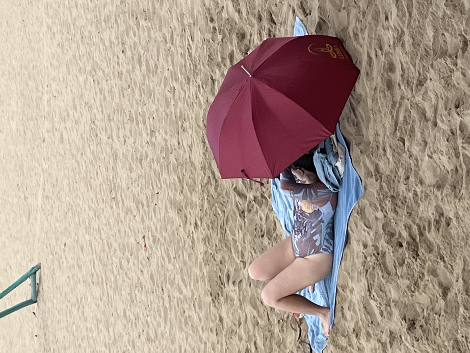 A person is lying on their back on a blue towel on a sandy beach. They are wearing a patterned swimsuit and have a red umbrella open over their head. The umbrella has a logo on it. In the background, part of a green structure is visible in the sand. The scene appears to be on a beach in Nha Trang, Vietnam.
