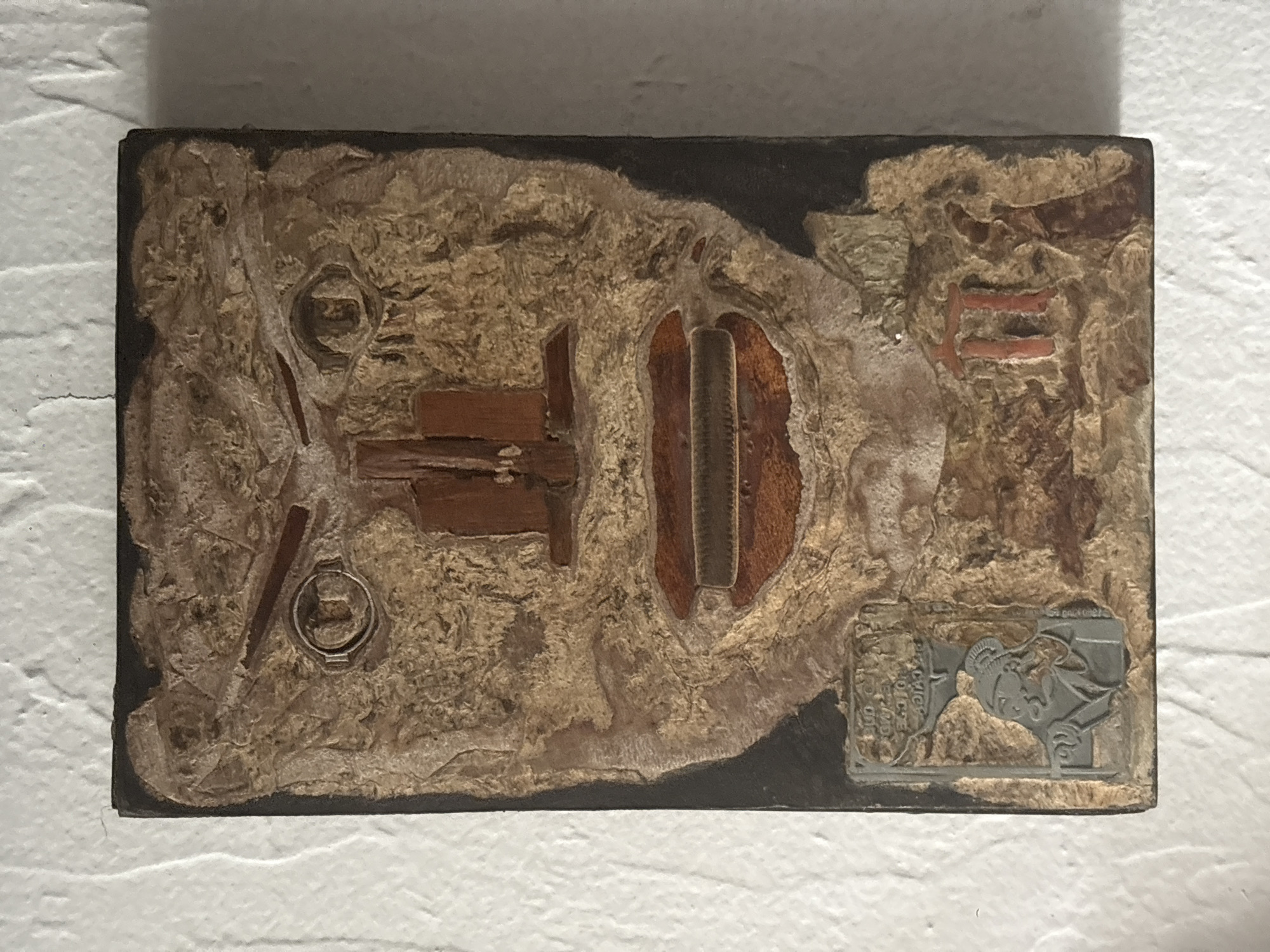The image shows a rectangular mixed-media artwork on a white, textured wall. The artwork features a dark brown or black background layer. The artwork's surface is uneven, with textured, beige and brown materials resembling a rocky or earthen landscape. Integrated within this texture are several embedded objects, including: Two circular metal rings with details inside them, arranged on the left side; a cross-shaped arrangement of small, reddish-brown wood fragments, and a central recessed area containing a vertical object with a ribbed texture. In the lower right corner, there is a small, gray metal emblem or plaque featuring a stylized image, possibly a figure or mythological creature. The artwork appears to be hanging on a wall. The location of this artwork in the city of Mexico City, Mexico is not knowable from this image.