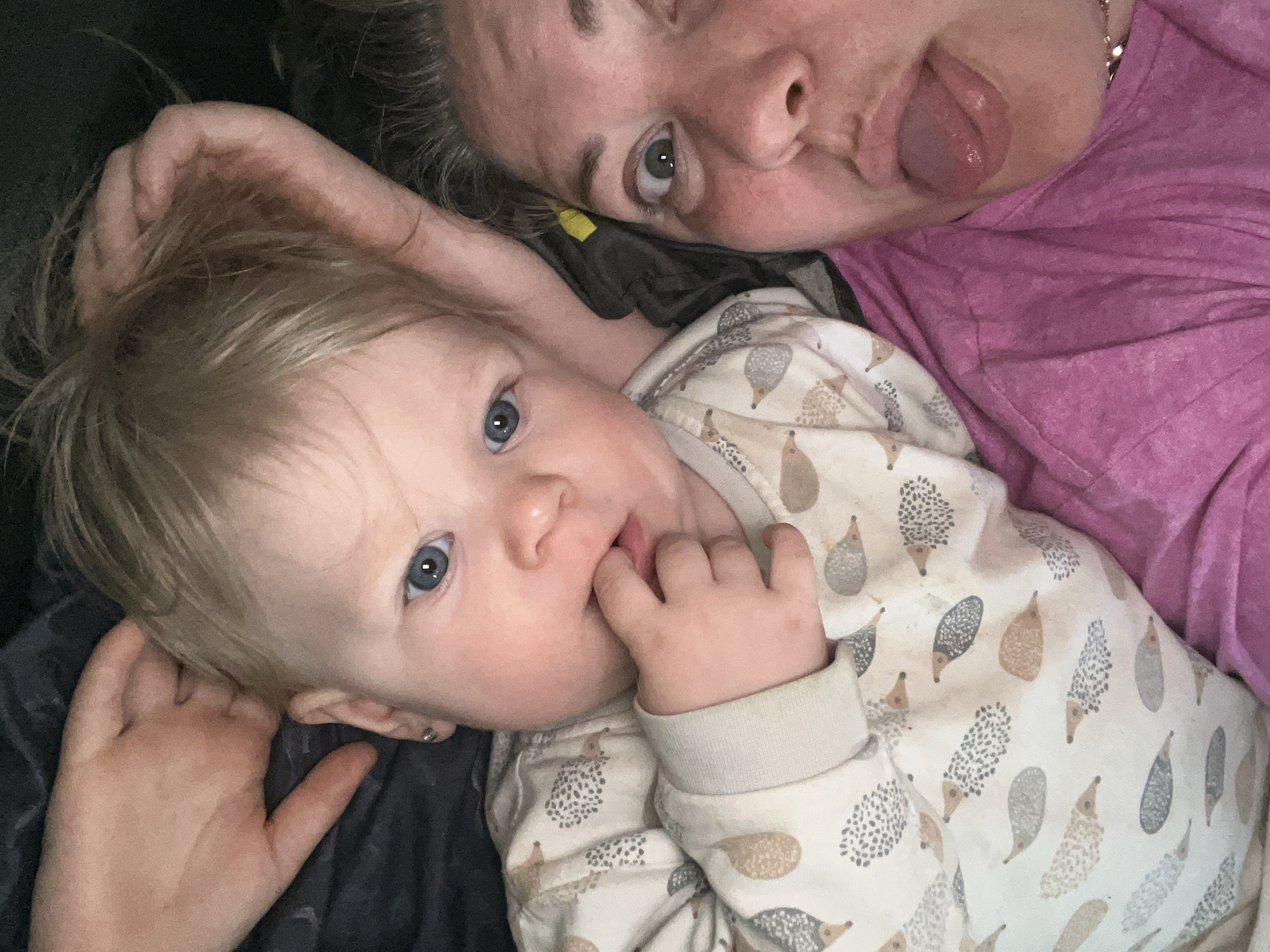 An overhead, close-up photograph captures an adult and a baby lying together, likely indoors in Фоки, Russia. The adult, positioned in the upper right, has fair skin, a grey/dark hairline, and is wearing a pink, textured top. She is playfully sticking her tongue out, with her right eye open and looking towards the camera. A silver chain is faintly visible near her neck. The baby, in the lower left, has fair skin, blonde hair, and striking blue eyes. They are wearing a light-colored onesie or pajamas adorned with a whimsical pattern of hedgehogs. The baby has fingers in their mouth and is also looking towards the camera with a curious expression. A dark fabric, possibly a blanket or clothing, is visible around the baby and behind the adult's head, suggesting a comfortable and relaxed setting, such as a bed or couch. The image conveys a moment of playful interaction and bonding between the two subjects. There is no visible text in the image.