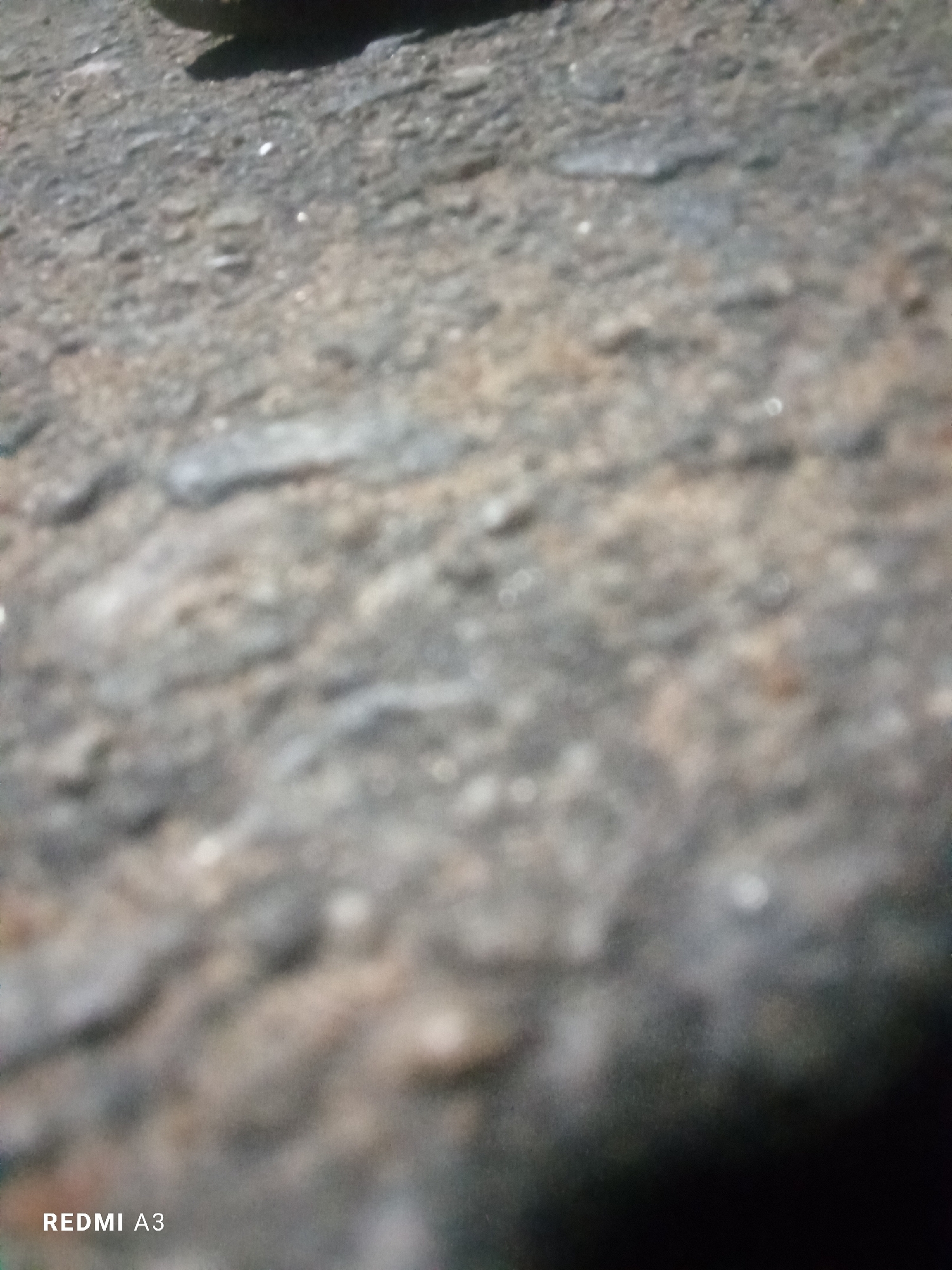 The image shows a close-up, out-of-focus view of a rough, textured surface, possibly concrete or asphalt, with some debris. In the upper left corner, a dark, curved shadow is visible, suggesting an object or a person casting it. The bottom left corner of the image displays the text "REDMI A3", indicating the device used to capture the photograph. There are no discernable landmarks or features that would allow for the identification of a specific location within Jalingo, Nigeria. The scene is devoid of any people, animals, or specific objects beyond the implied shadow and the text overlay.