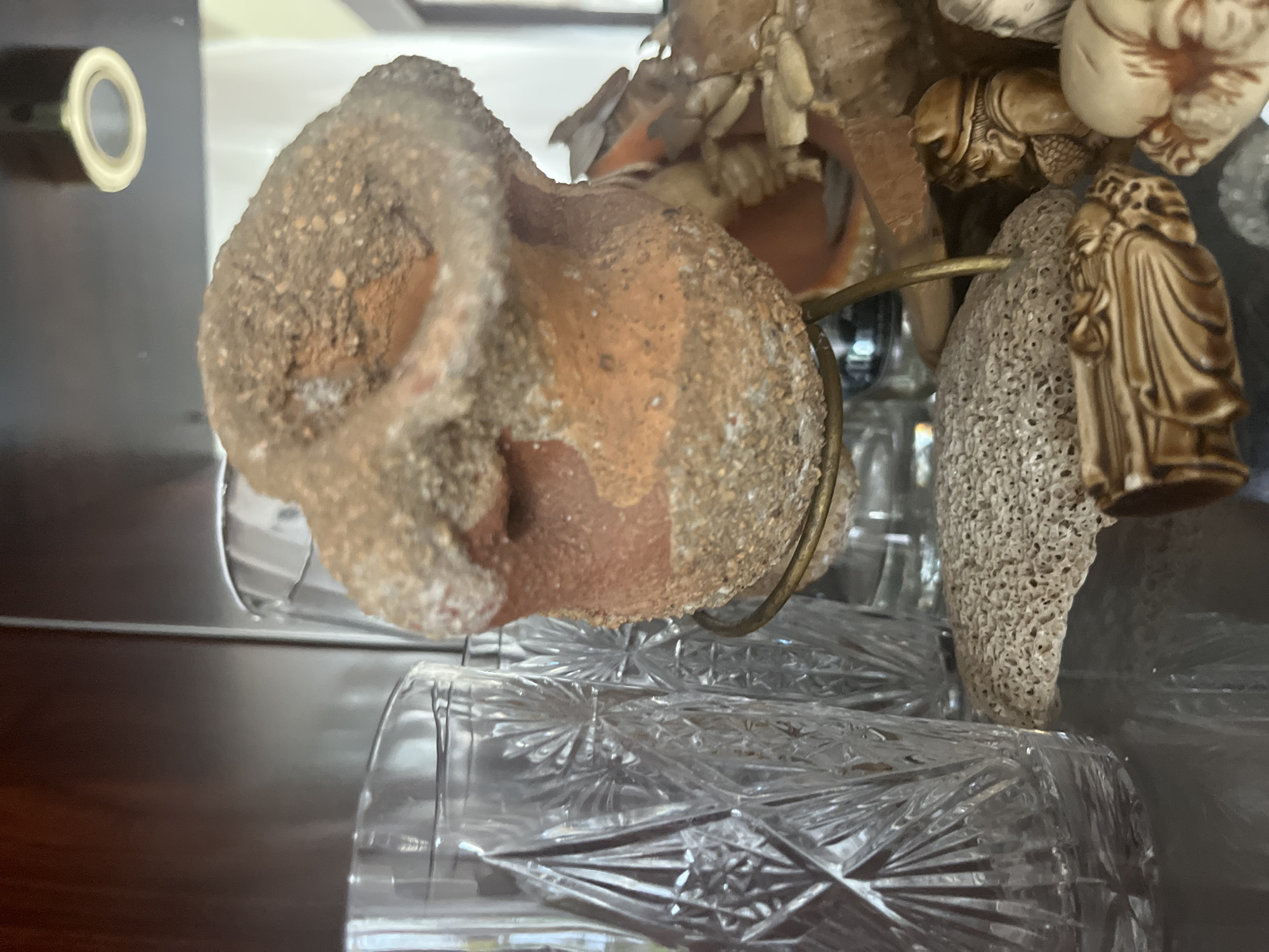 A close-up shot reveals a collection of various decorative objects, likely displayed on shelves in a home. In the foreground, a roughly textured, reddish-brown ceramic pot sits on a thin, brass-colored stand. The pot appears old and weathered, with visible grains of sand or grit embedded in its surface. To the left of the pot, a tall, clear glass with an intricate, faceted pattern stands prominently. Behind the pot, partially obscured, is another clear glass, simpler in design. Below these items, a light-colored, porous-looking object, possibly a piece of coral or pumice, lies on the reflective surface of the shelf. To the right and in the background, other decorative elements are visible, including small, carved figures made of a light-colored material, some appearing to be people. The overall impression is that of a curated display of trinkets and curiosities. The scene is set indoors, likely in a room with dark wood shelving. No specific location in Vladimirovka, Russia, can be identified from the image.