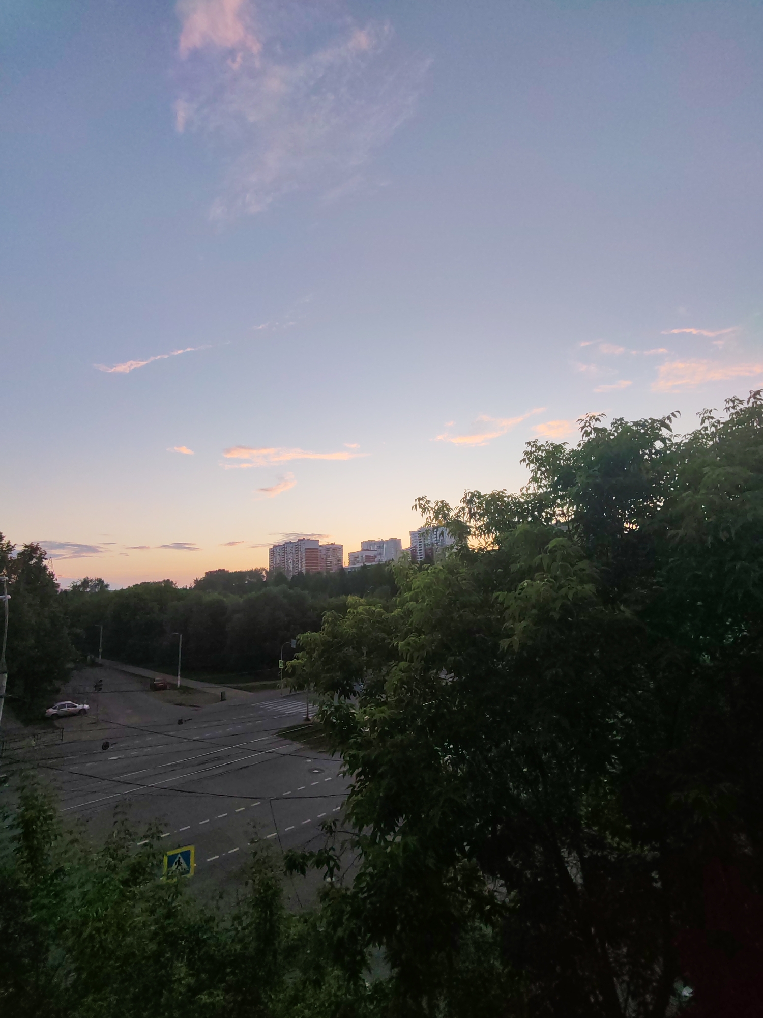 The image captures a panoramic view of a city street at dusk, likely in Izhevsk, Russia. The sky is a soft gradient of pale blue transitioning to a warm orange and pink horizon, with wispy clouds scattered across it. In the background, a line of modern apartment buildings stands against the horizon, hinting at a residential area. A dense band of dark green trees separates the buildings from the foreground.
The foreground is dominated by a street intersection with traffic lights and lane markings. Several vehicles, including a white car and a red car, are visible on the asphalt. A pedestrian crossing is marked by white stripes. A yellow pedestrian crossing sign with a blue triangle and a white silhouette of a person is posted on the side of the road. The street appears to be relatively quiet, with no pedestrians visible. Large trees with dark green foliage frame the right side of the image, obscuring parts of the scene and adding depth. The overall impression is one of a peaceful urban evening, with nature and city infrastructure coexisting.