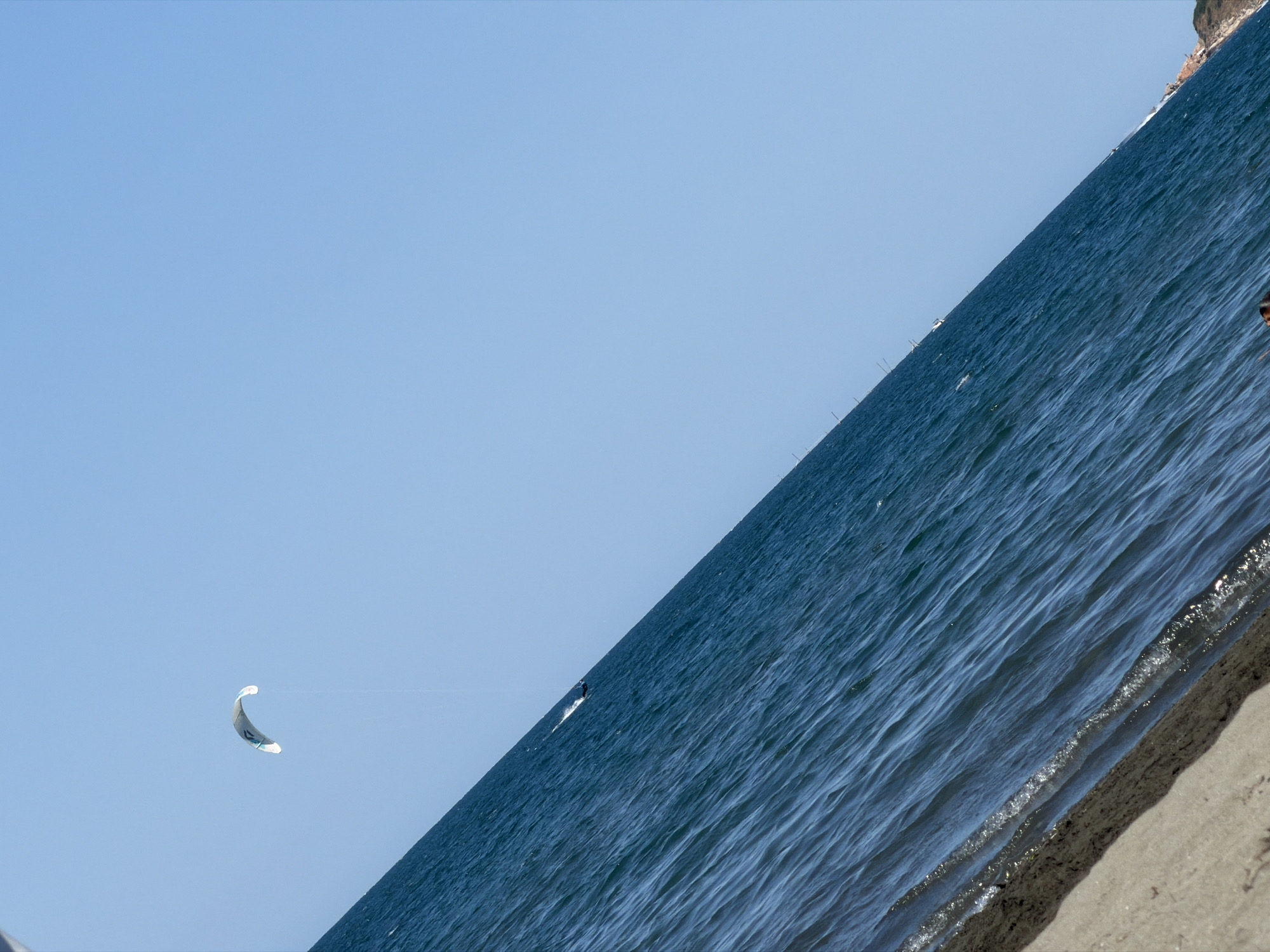 The image depicts a wide shot of the sea under a clear blue sky, taken from a beach. The perspective is tilted, with the shoreline running diagonally from the bottom left to the middle right of the frame. The water is a deep blue, with small white-capped waves indicating a gentle breeze. A white kite is visible in the upper left quadrant of the sky, attached by thin lines to a kite surfer who is actively riding the waves further down the sea. A sandy beach occupies the bottom portion of the image. The location is identified as the city of Atiya, Bulgaria.