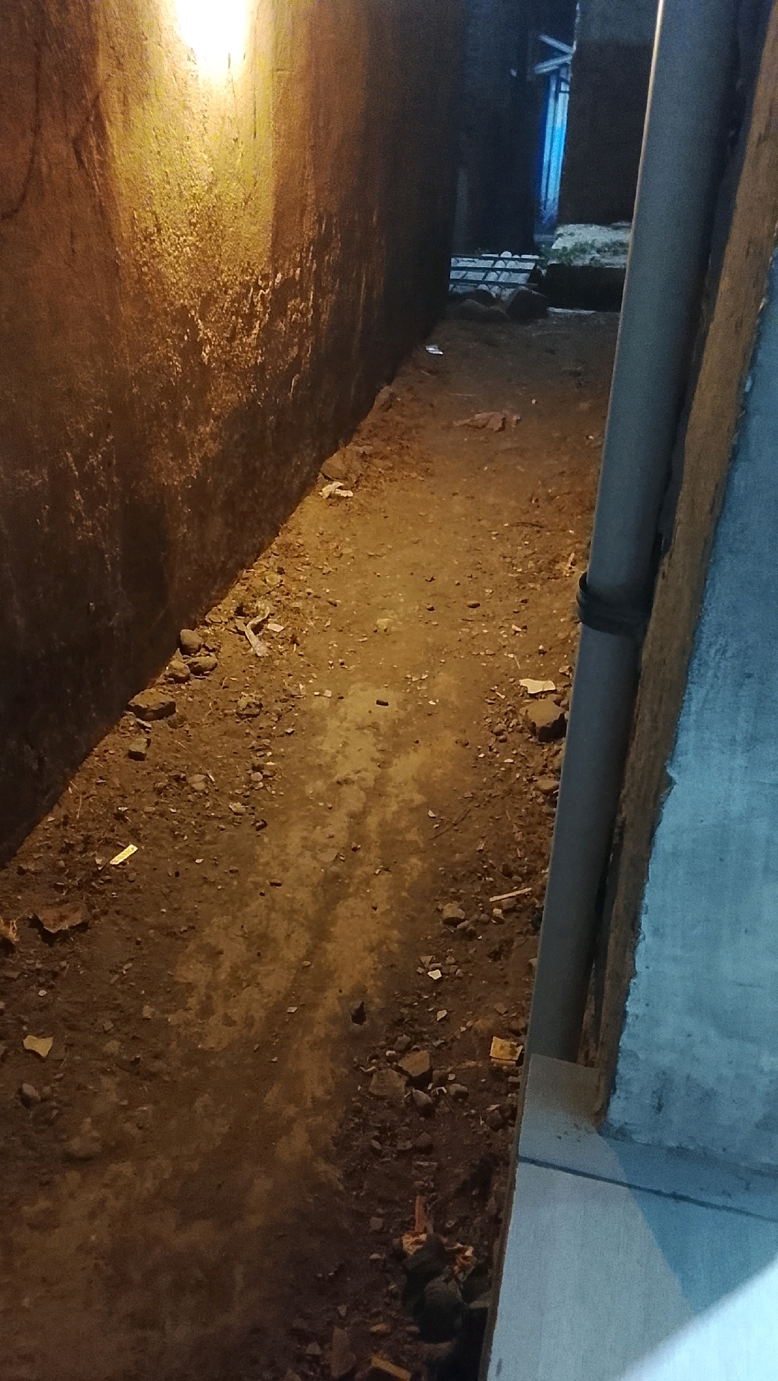 The image shows a narrow alleyway at night in Ujungrusi, Indonesia. The ground is unpaved and covered with dirt, small stones, and scattered debris. On the left, a rough, textured wall, illuminated by a warm light source from above, casts a shadow. On the right, a grey pipe runs vertically along a concrete structure. In the background, there is a glimpse of what appears to be a doorway or opening with blue light. The scene suggests an outdoor, utilitarian space, possibly a service passage or back alley.