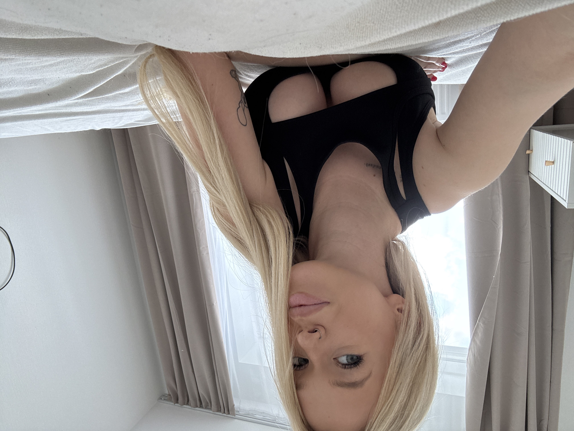 A blonde woman with blue eyes and a nose ring is lying on a white bed, looking towards the camera. She is wearing a black, cut-out tank top. Her hair is long and blonde, falling over her shoulders. To her left is a white bedside table with wooden knobs, and behind her are light brown curtains. The room appears to be a bedroom. The location is Petro-Slavyanka, Russia.