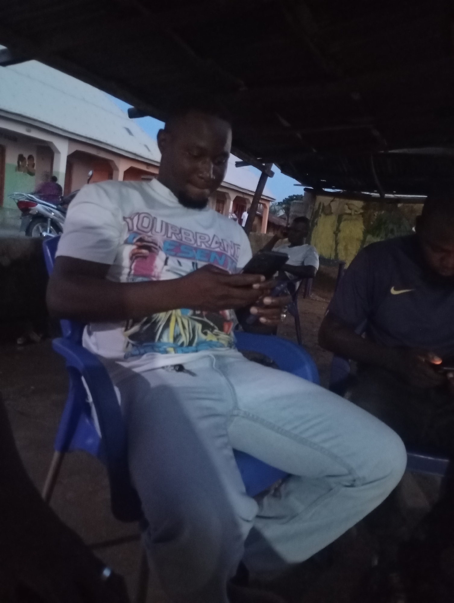 The image depicts an outdoor scene at dusk in Shendam, Nigeria. In the foreground, a man is seated in a blue plastic chair, engrossed in his smartphone. He wears a white t-shirt with a graphic design and light-colored pants. To his right, another man is also seated and looking at his phone, wearing a dark blue t-shirt with a yellow Nike logo. In the background, other individuals are visible, along with buildings and a motorcycle. The environment appears to be a simple outdoor gathering area, possibly a roadside stall or a communal space, with a corrugated metal roof providing shelter. The lighting suggests it is either early morning or late evening.